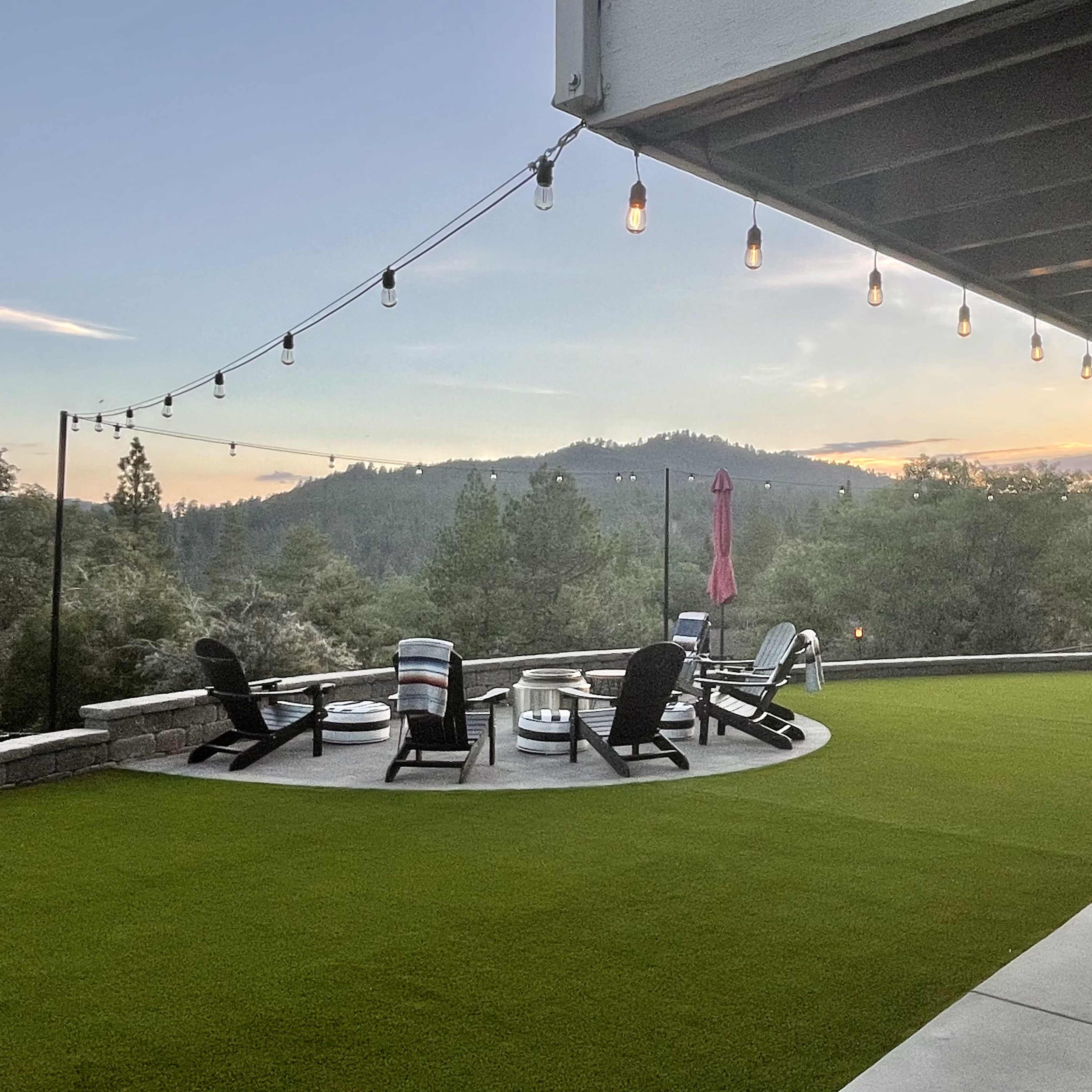 A spacious patio with circular seating, firepits, and string lights overlooks a mountainous landscape at sunset.