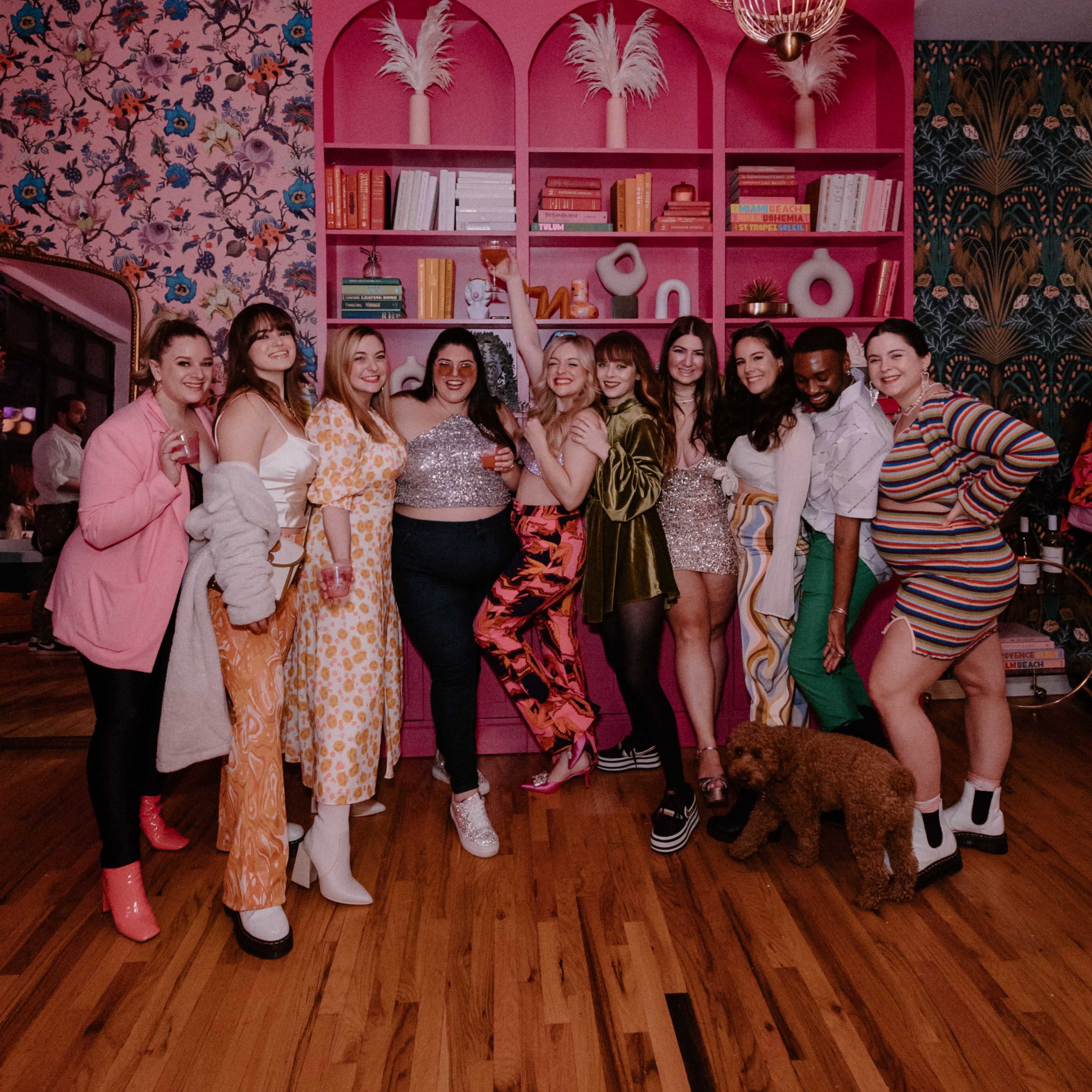 A group of women pose together in a colorful room adorned with floral wallpaper and a vibrant pink bookshelf.