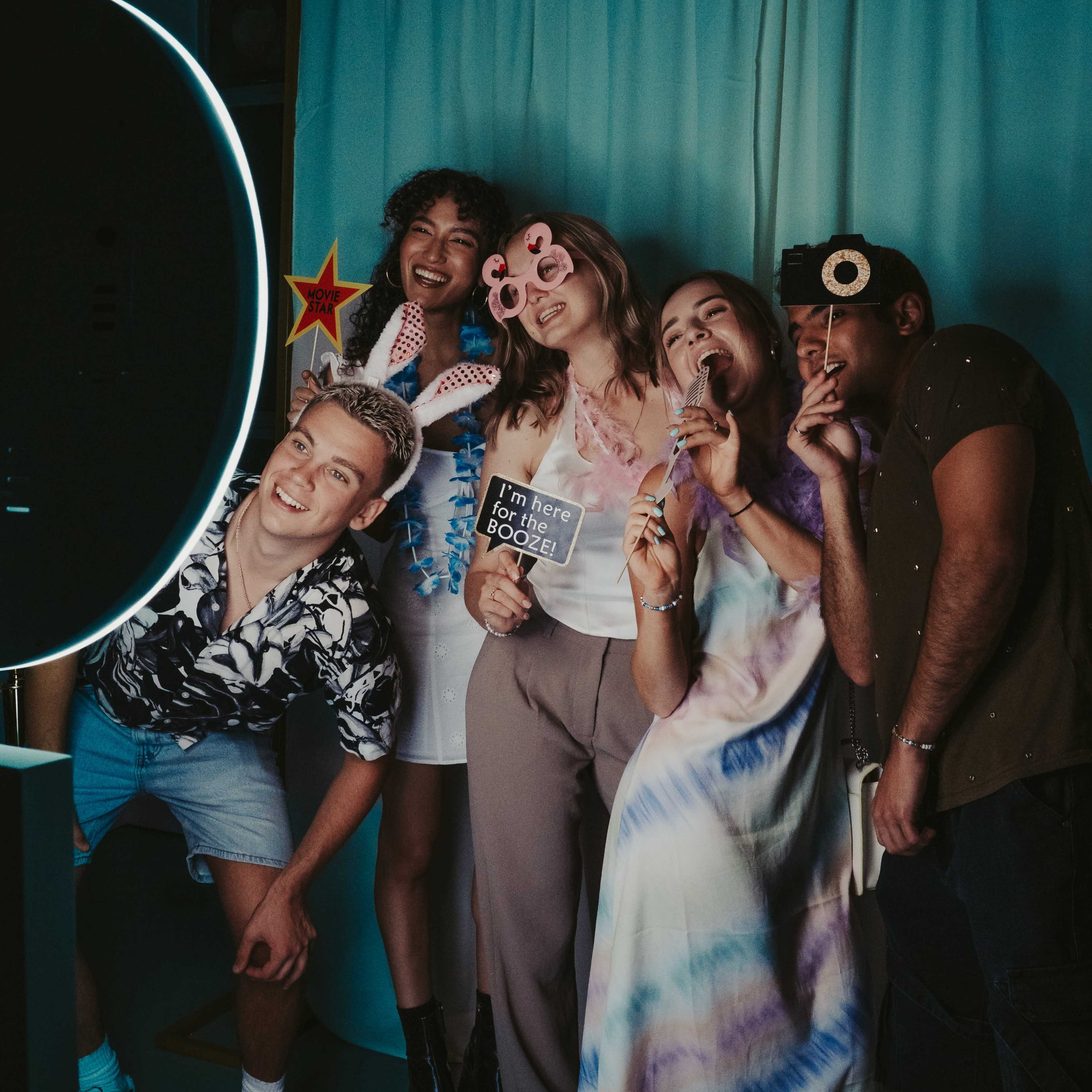 A group of five friends poses playfully in front of a circular light, holding props and making fun gestures against a backdrop of blue fabric.