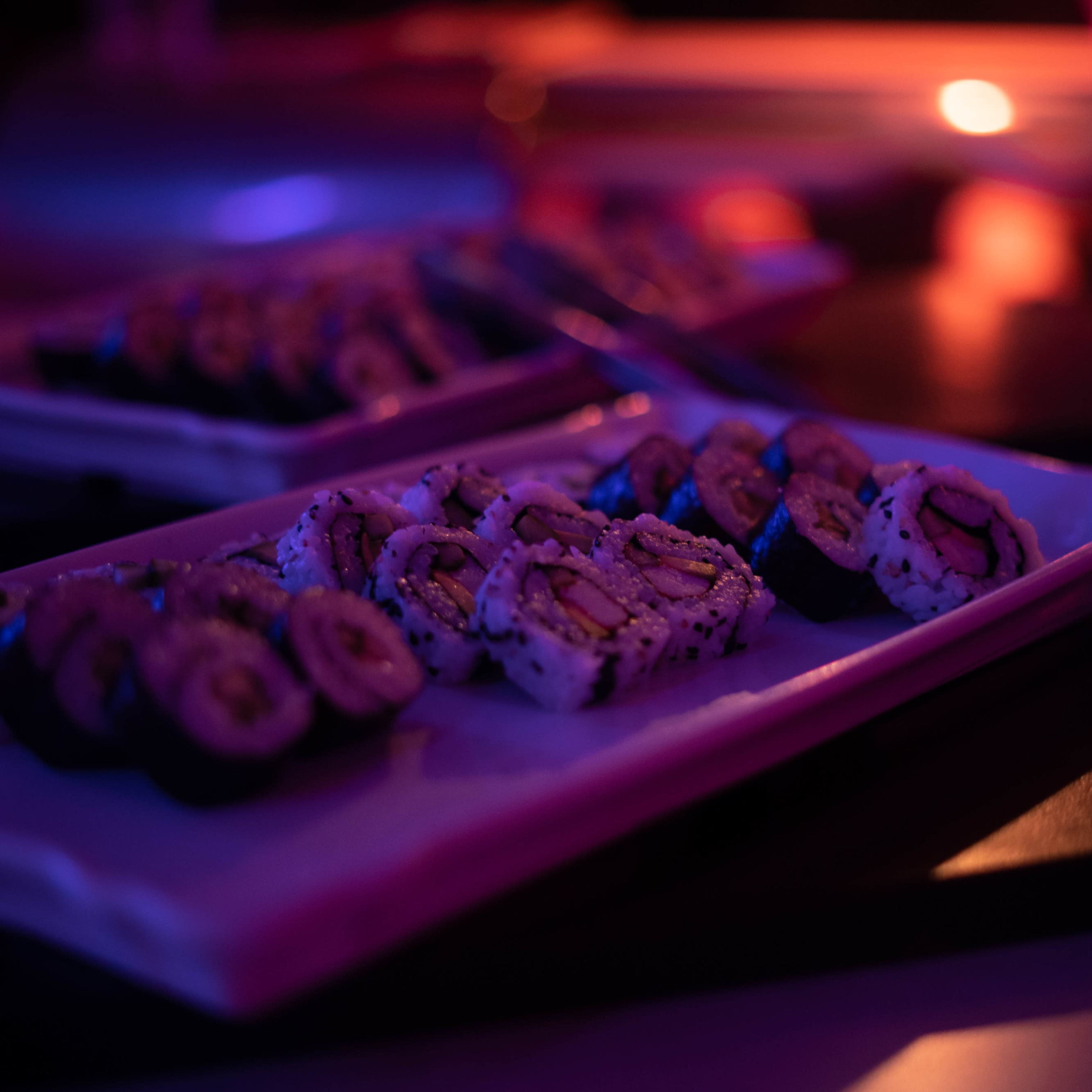 Sushi rolls are arranged on a white plate, illuminated by soft purple and orange lighting.