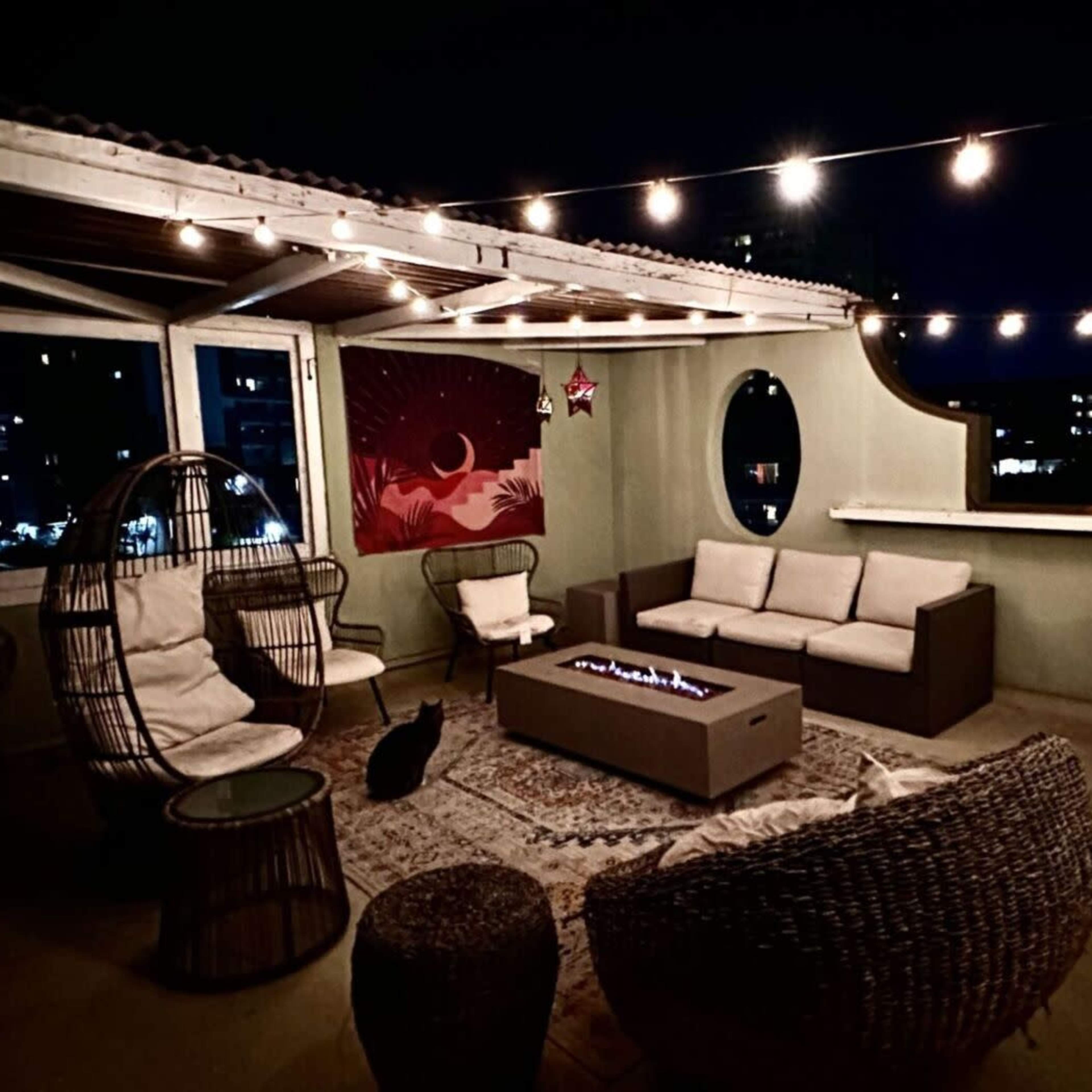 A rooftop patio features a fire pit surrounded by seating, decorative string lights overhead, and a cat resting on the floor.