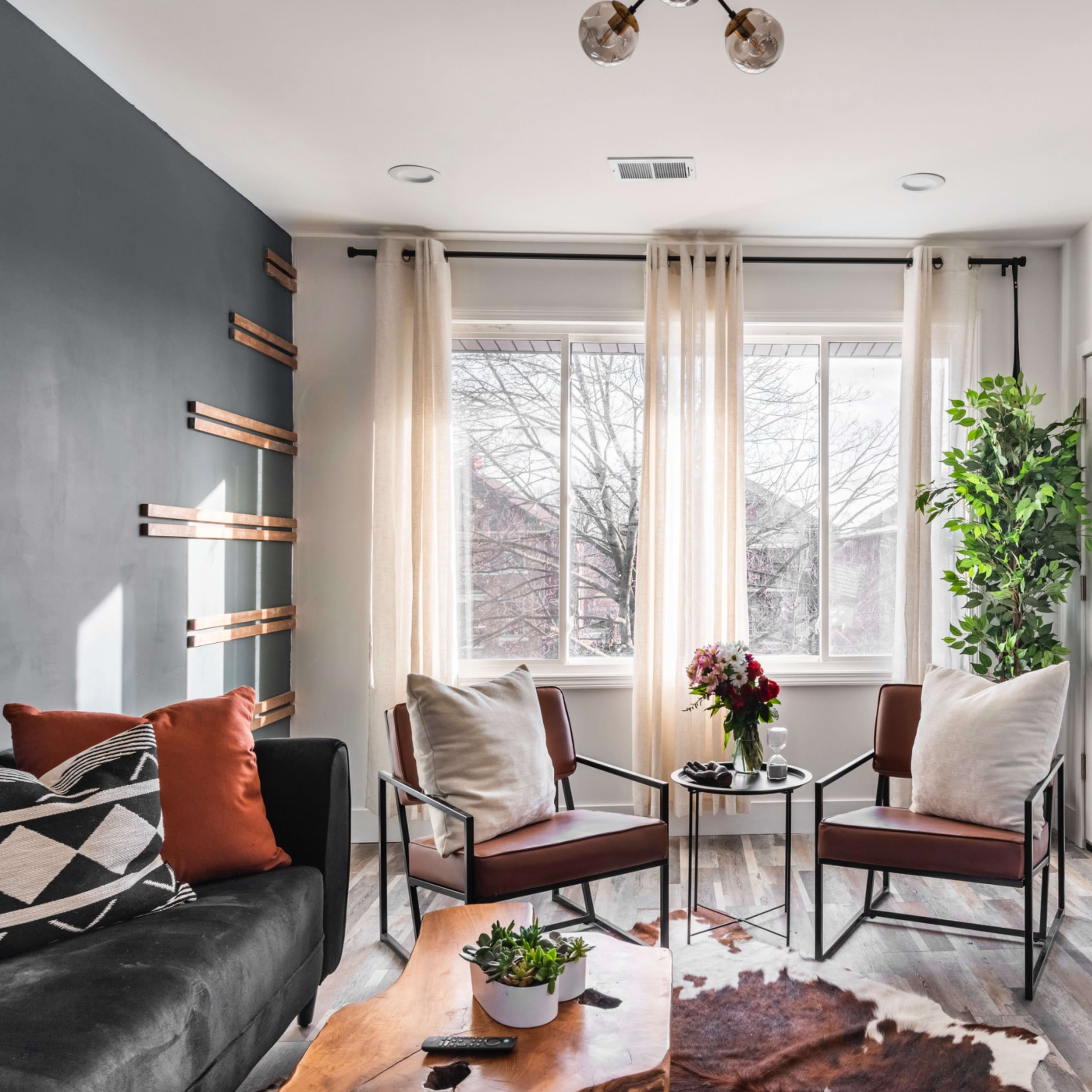 A modern living room featuring a dark accent wall, two chairs with cushions, a coffee table, and large windows with sheer curtains allowing natural light to fill the space.