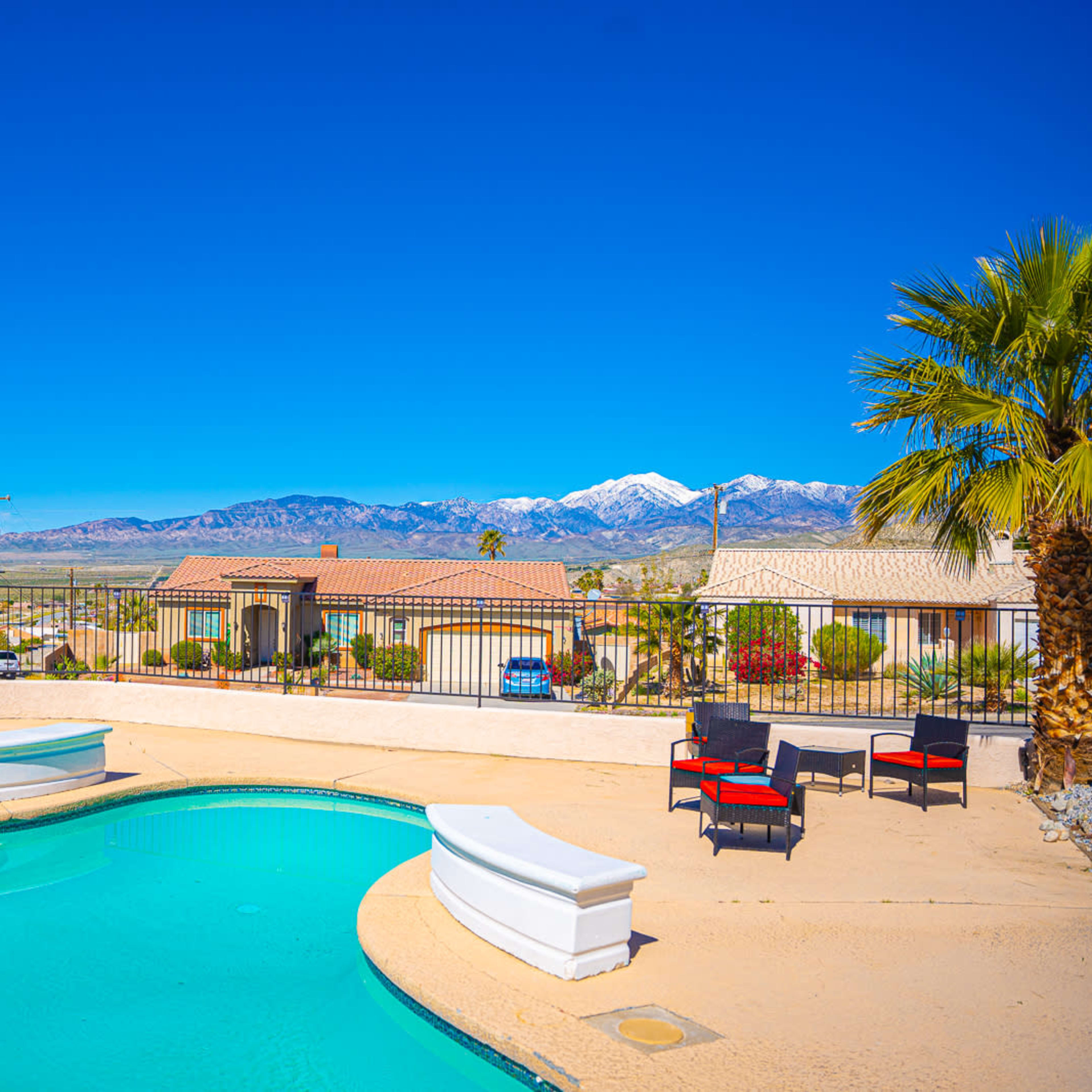 A swimming pool surrounded by lounge chairs is situated on a patio, with mountains and blue skies in the background.