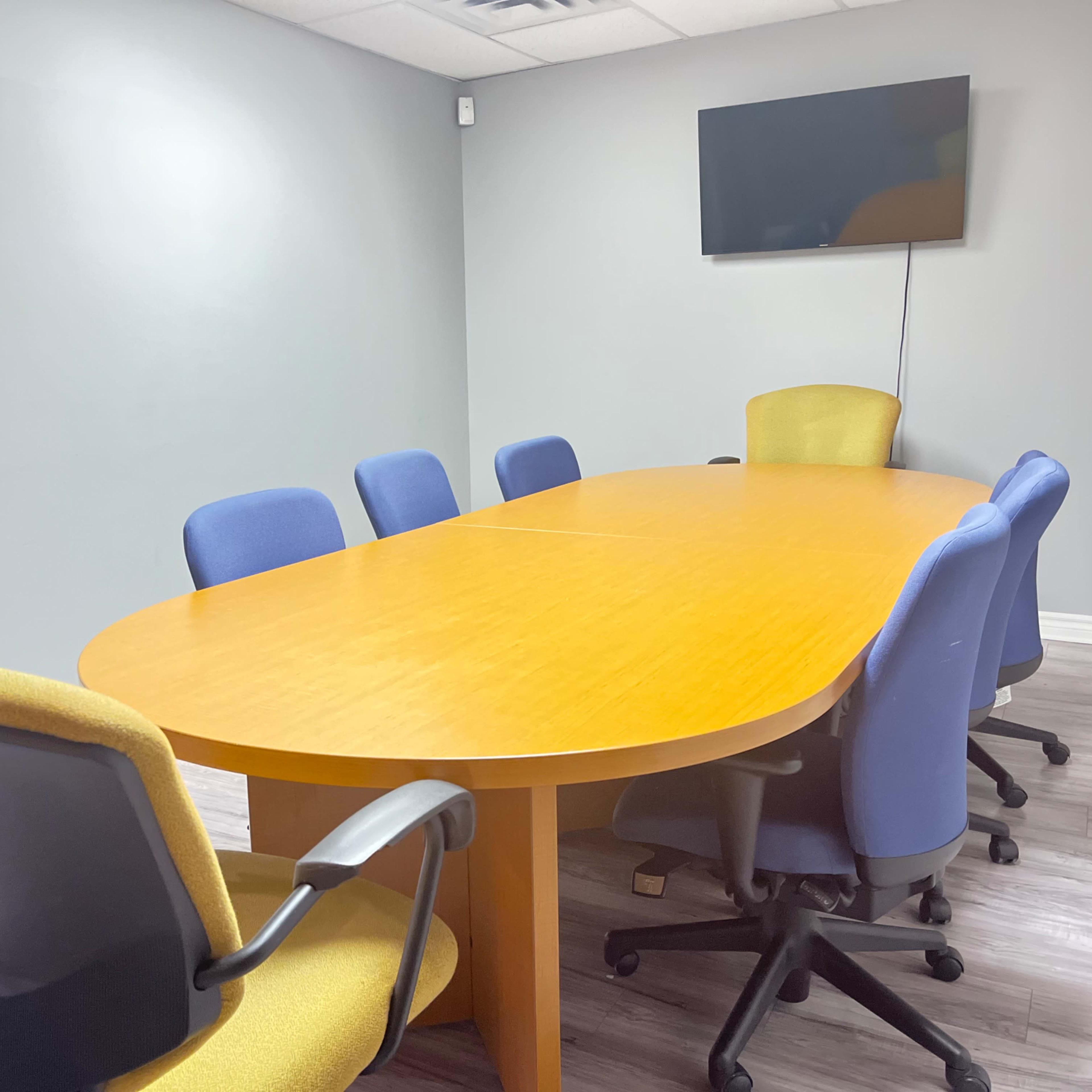 A conference room features a large oval wooden table surrounded by eight office chairs, with a wall-mounted television in the background.