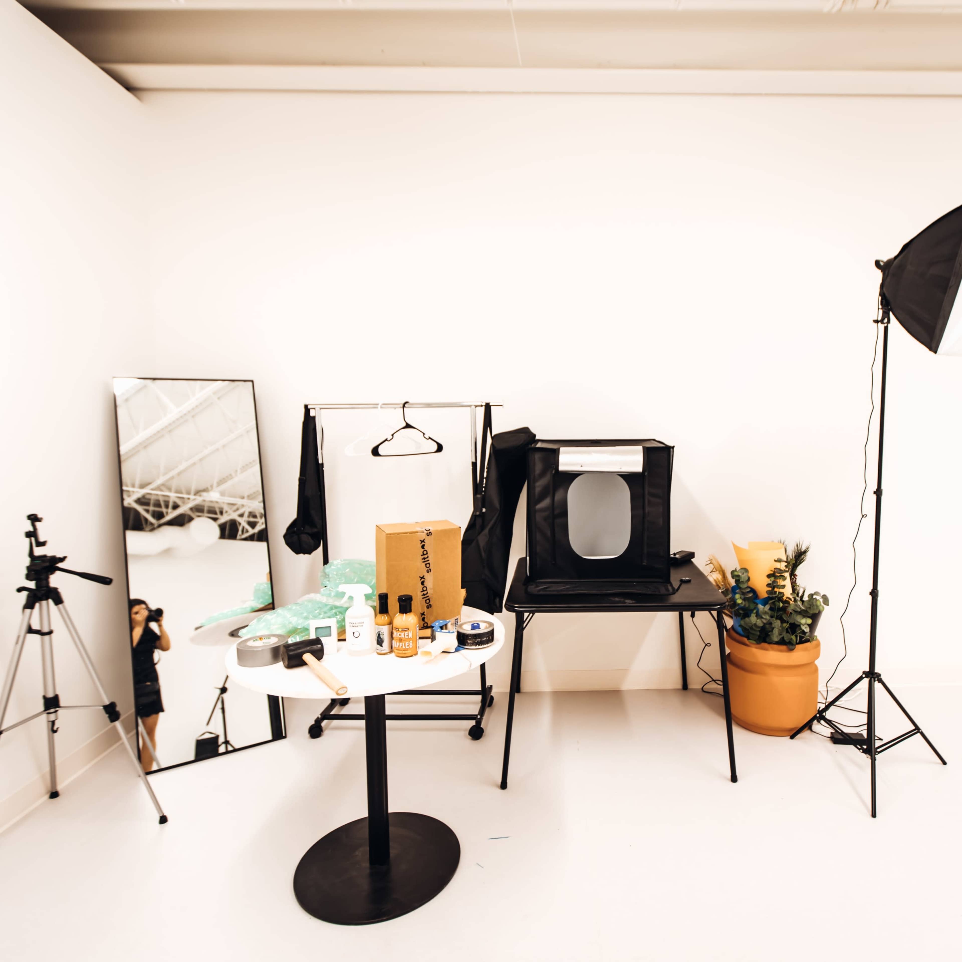 A small studio space features a table with various beauty products, a tripod, a mirror, a chair with a portable backdrop, and a potted plant.