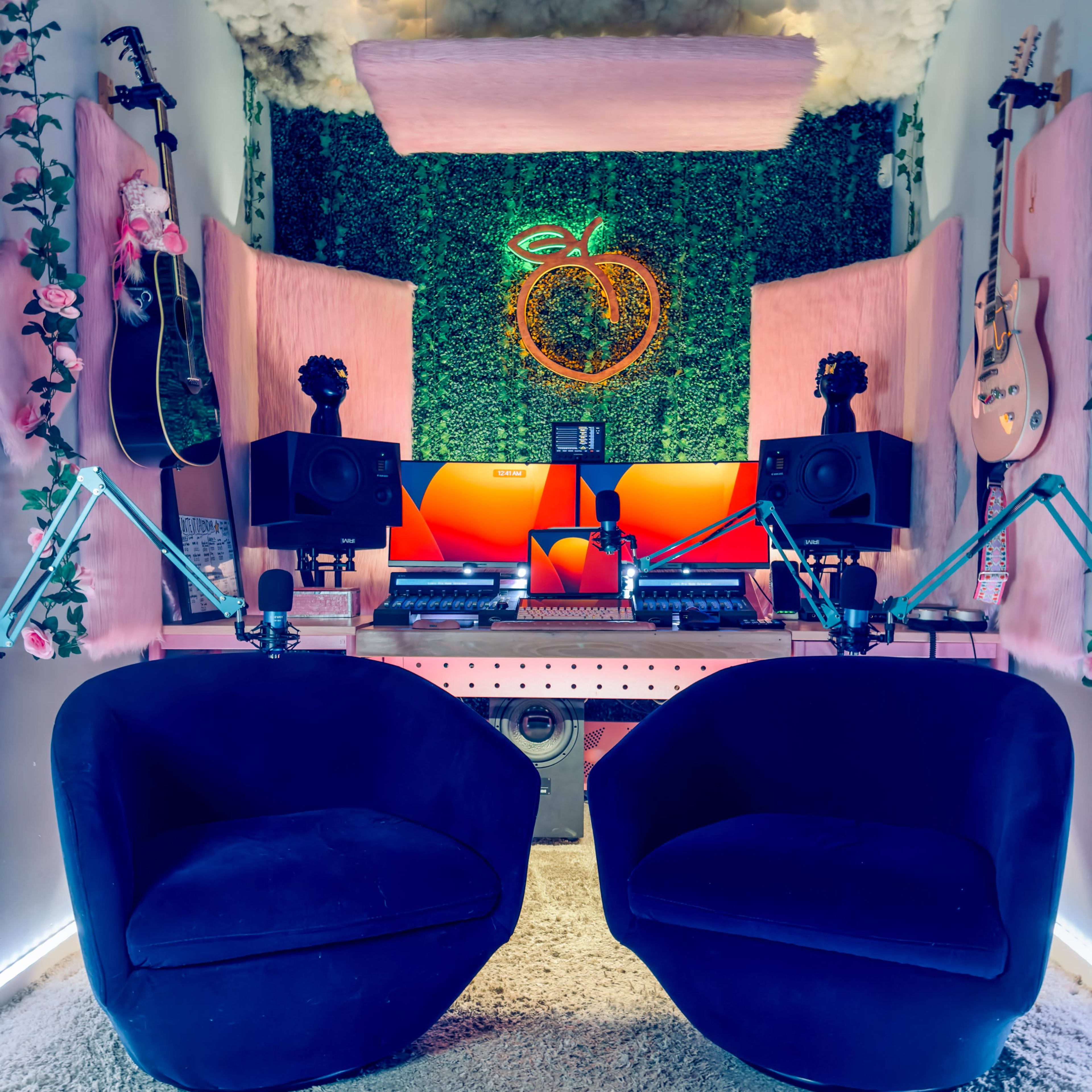 A small, cozy recording studio featuring two black chairs, musical instruments, sound equipment, and vibrant green wall decor with artificial foliage.