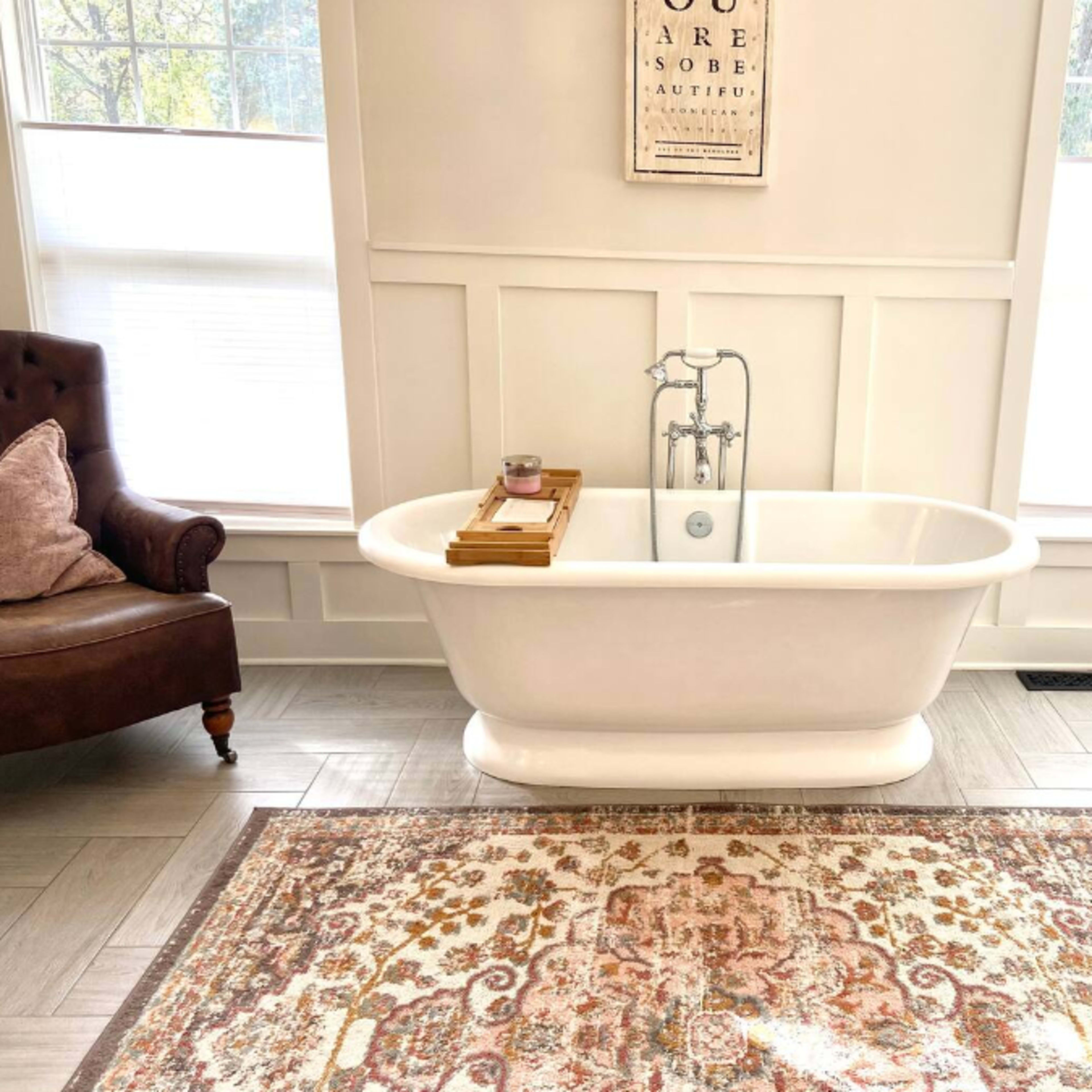 A modern bathroom featuring a freestanding white bathtub, a plush armchair, a decorative rug, and a wooden accent table.