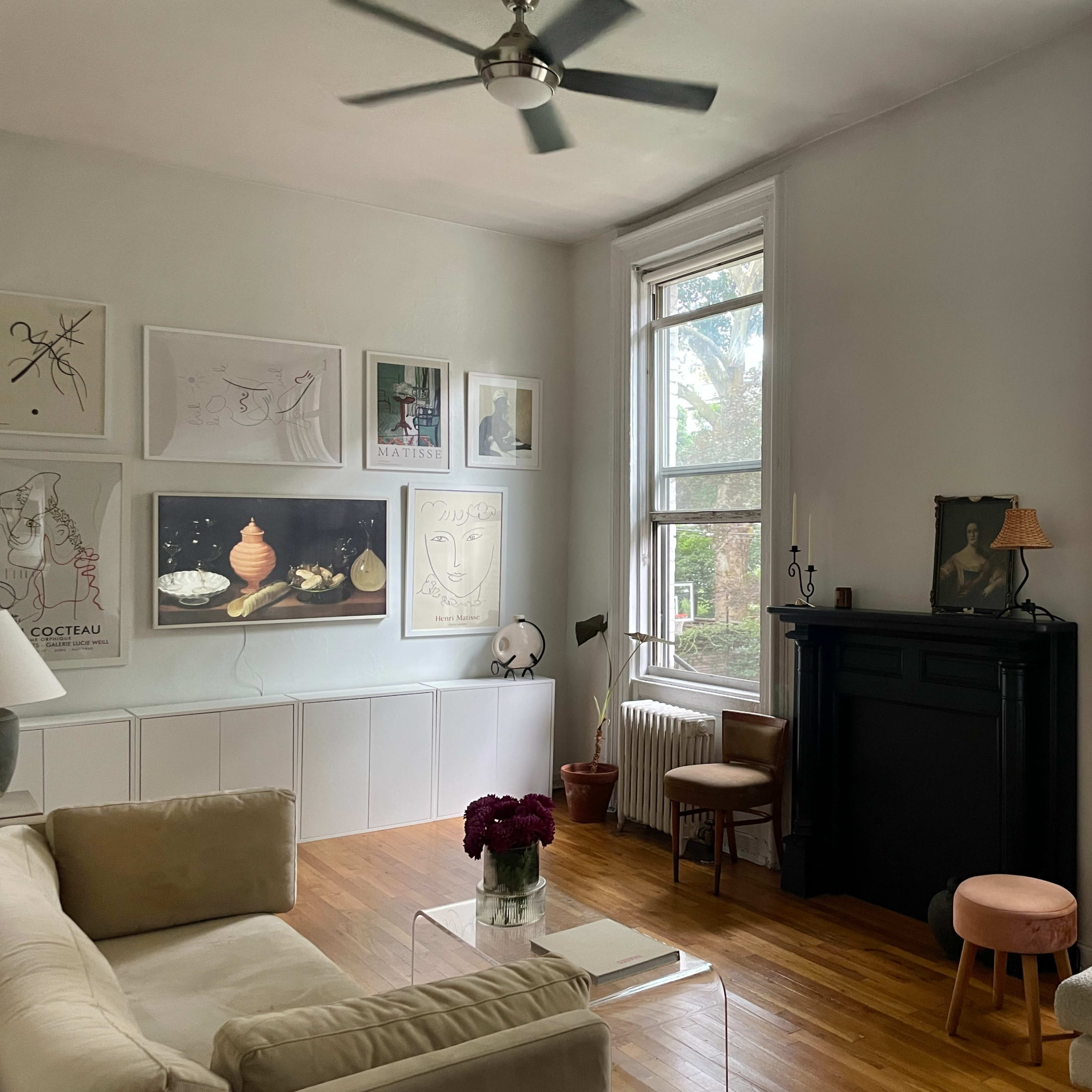 The room features a light-colored sofa, a glass coffee table, and a decorative fireplace, with large windows allowing natural light and views of greenery outside.