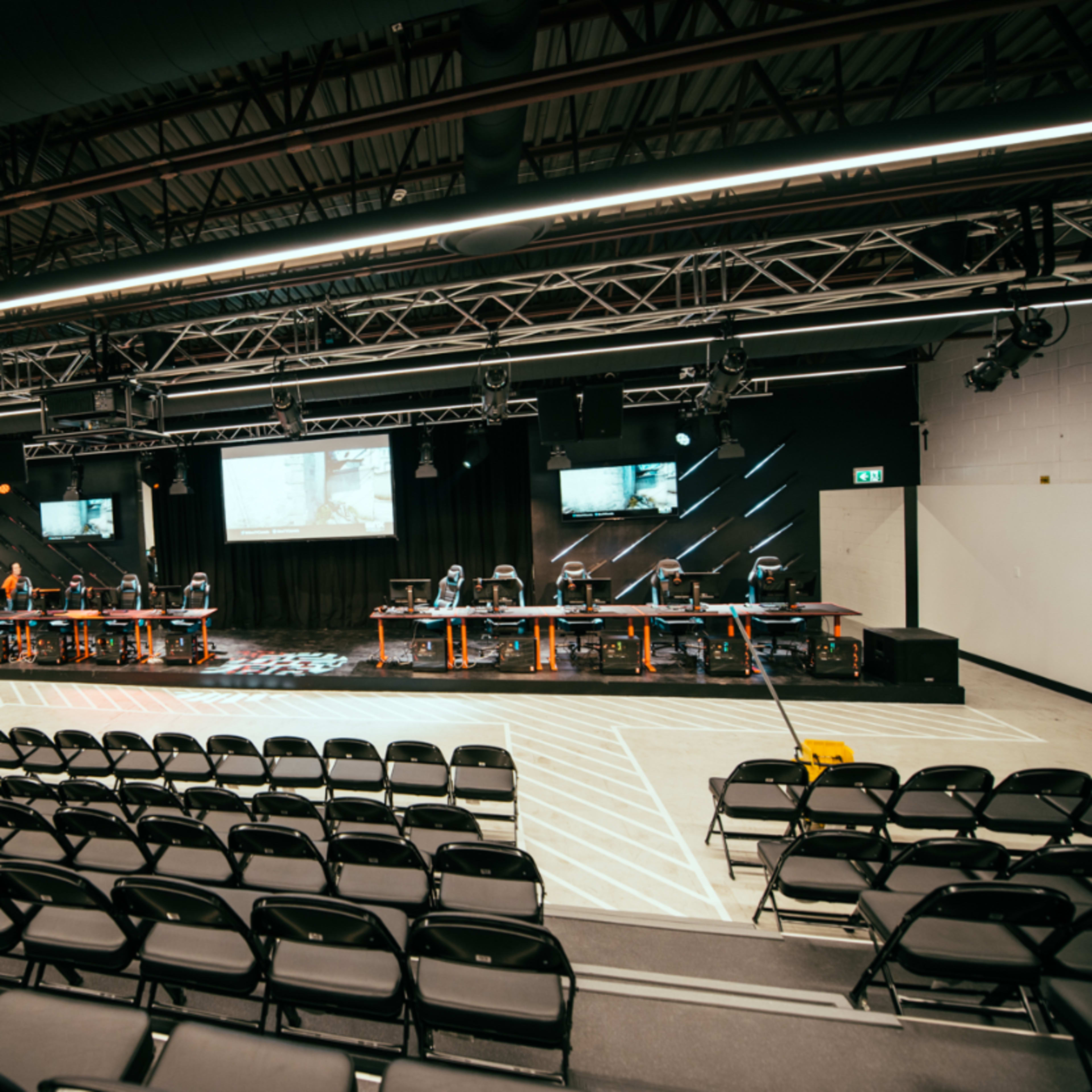 An empty auditorium features rows of black chairs facing a stage with multiple computer stations and screens.