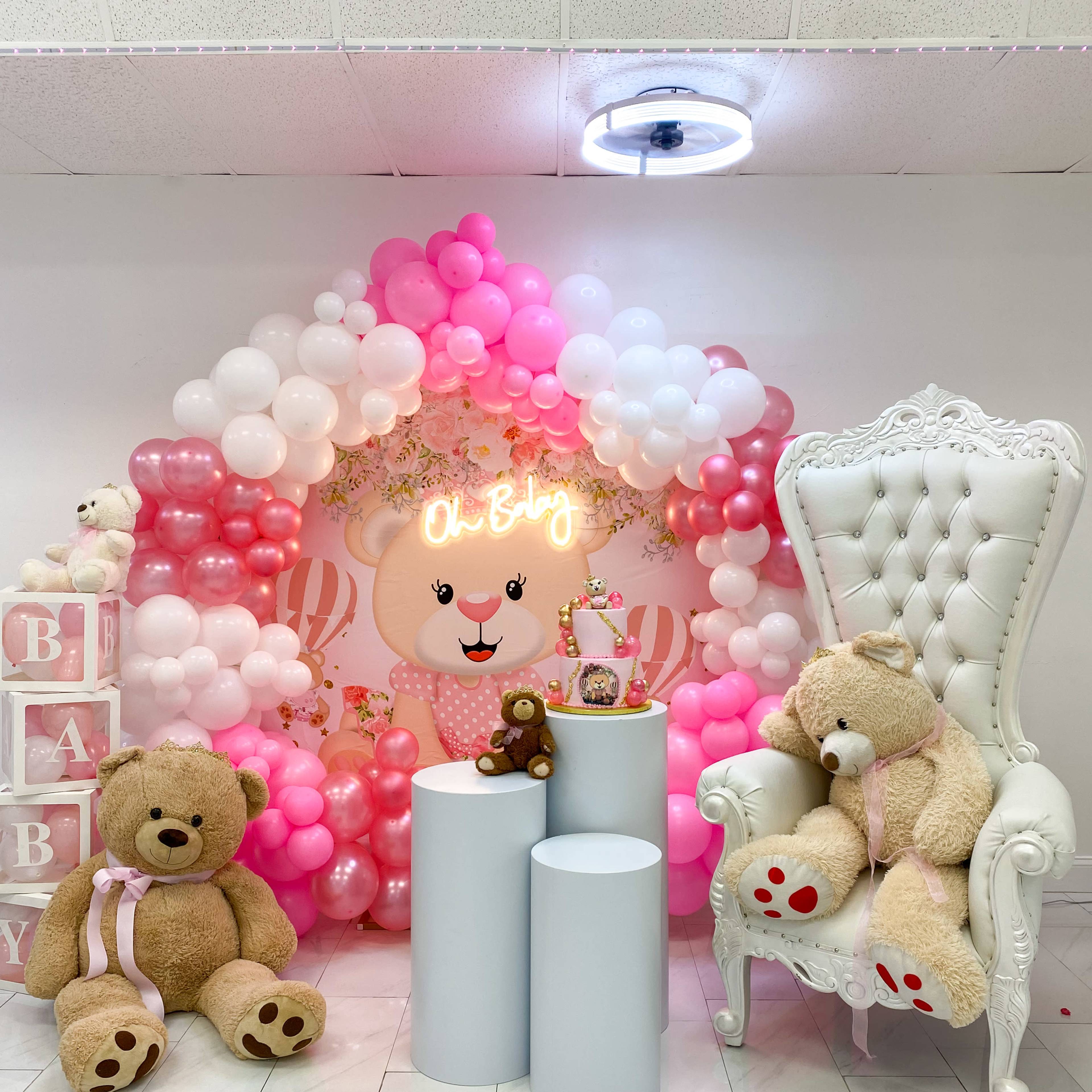 A decorative setup featuring a backdrop of pink and white balloons, a glowing "Oh Baby" sign, plush teddy bears, and a large white chair.