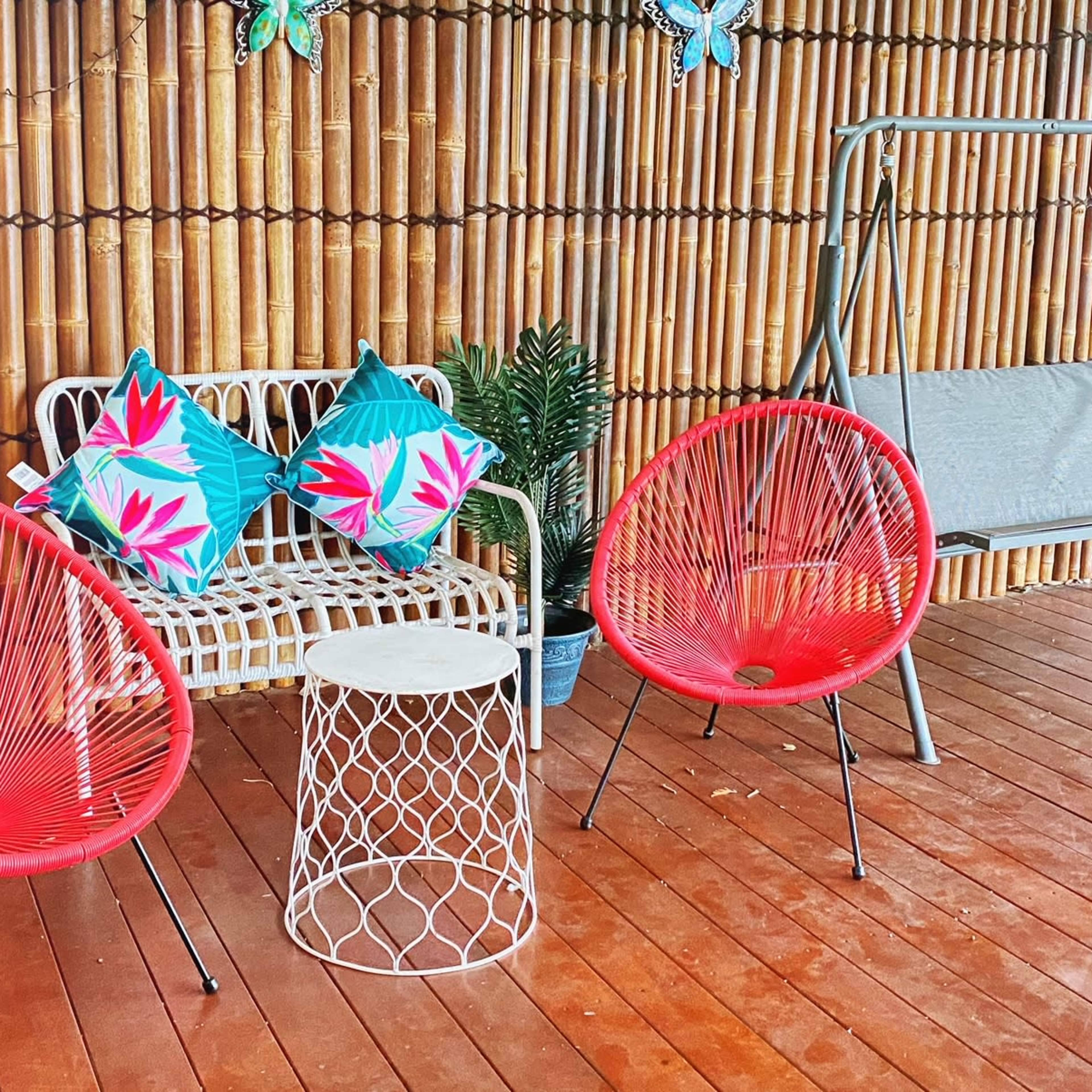 A cozy outdoor seating area with two red chairs, a small white table, a swing bench, and a bamboo backdrop.