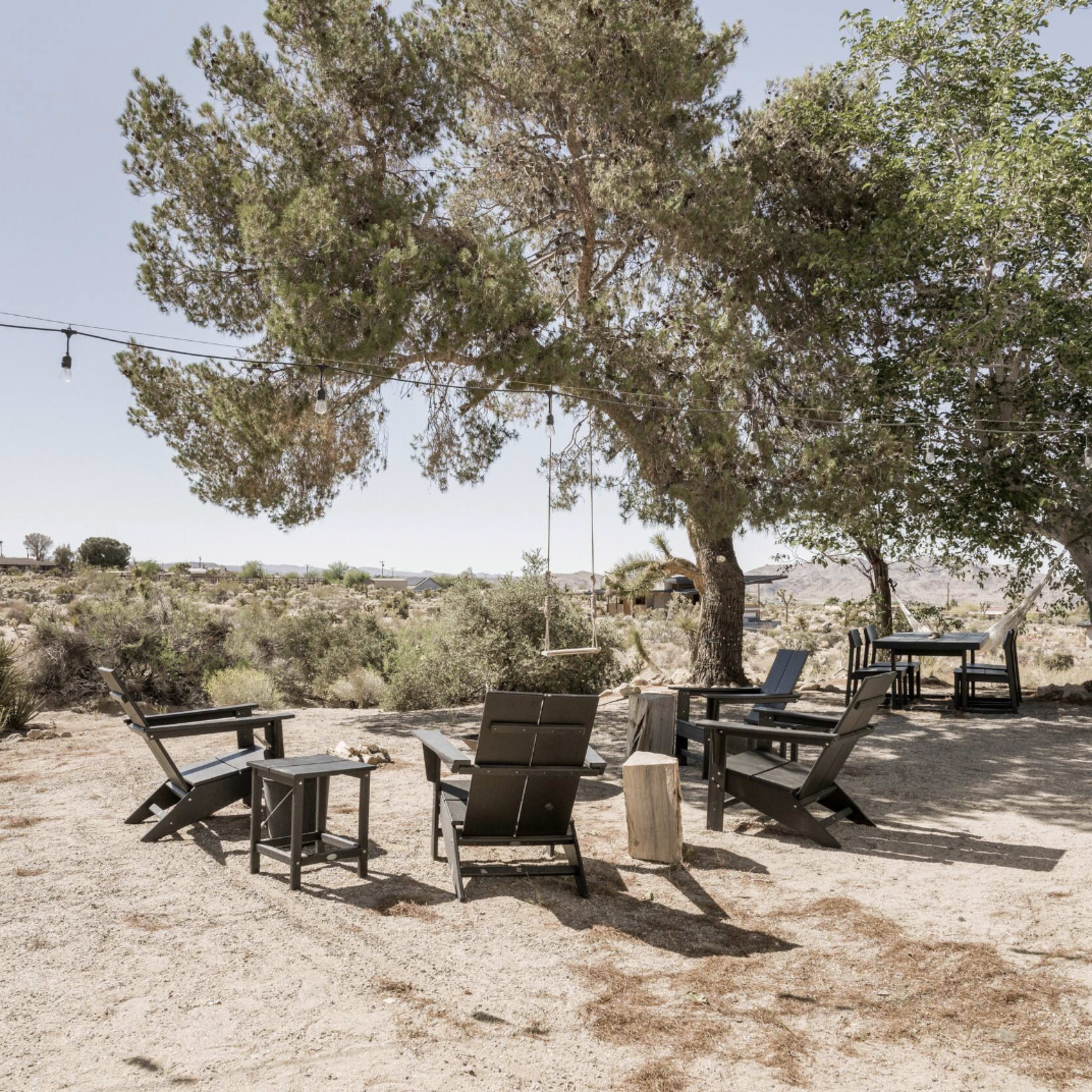 A seating area with black outdoor chairs and tables under two trees in a desert landscape.