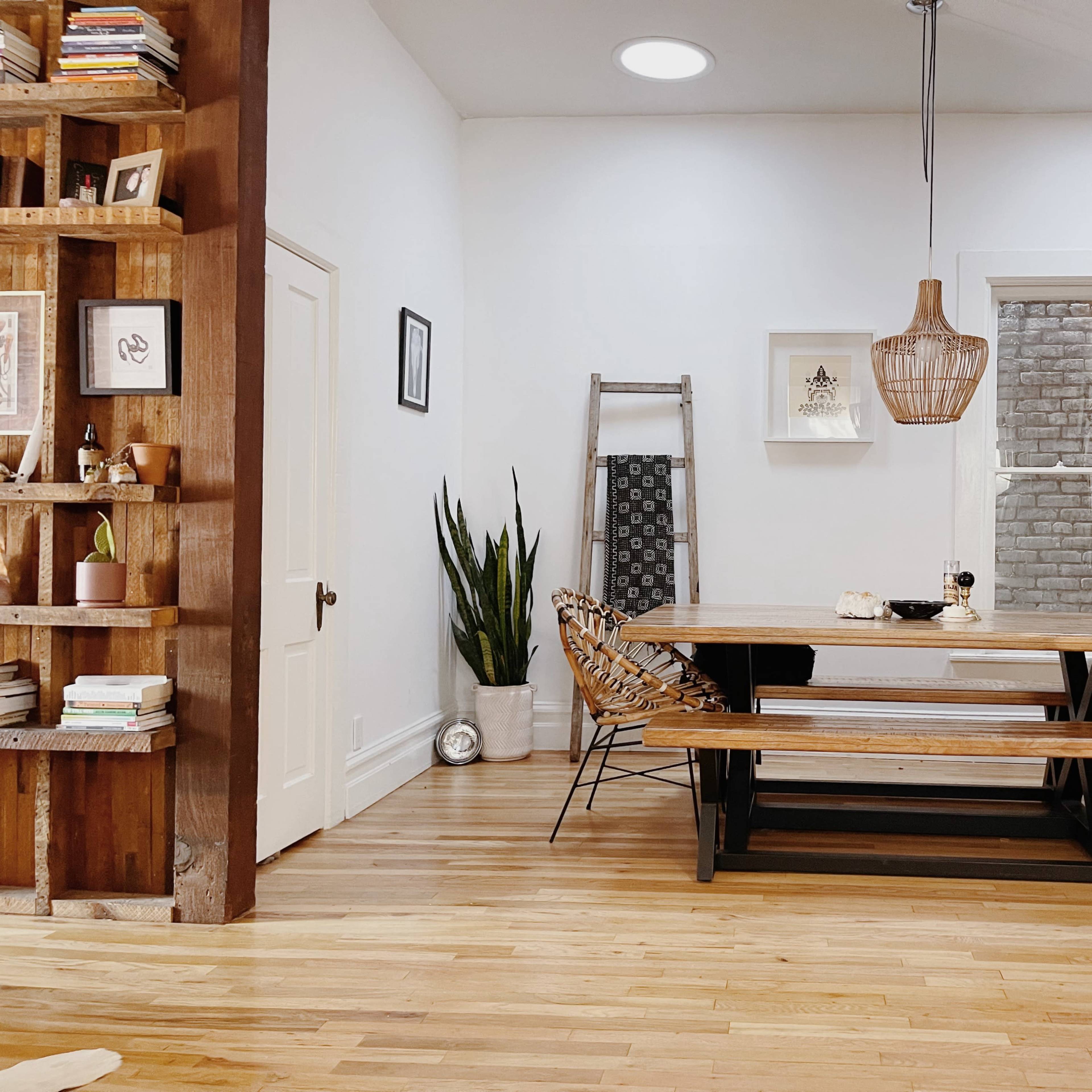 The scene shows a modern dining area featuring a wooden table surrounded by wicker chairs, a ladder leaning against a wall, and a large mirror reflecting natural light.