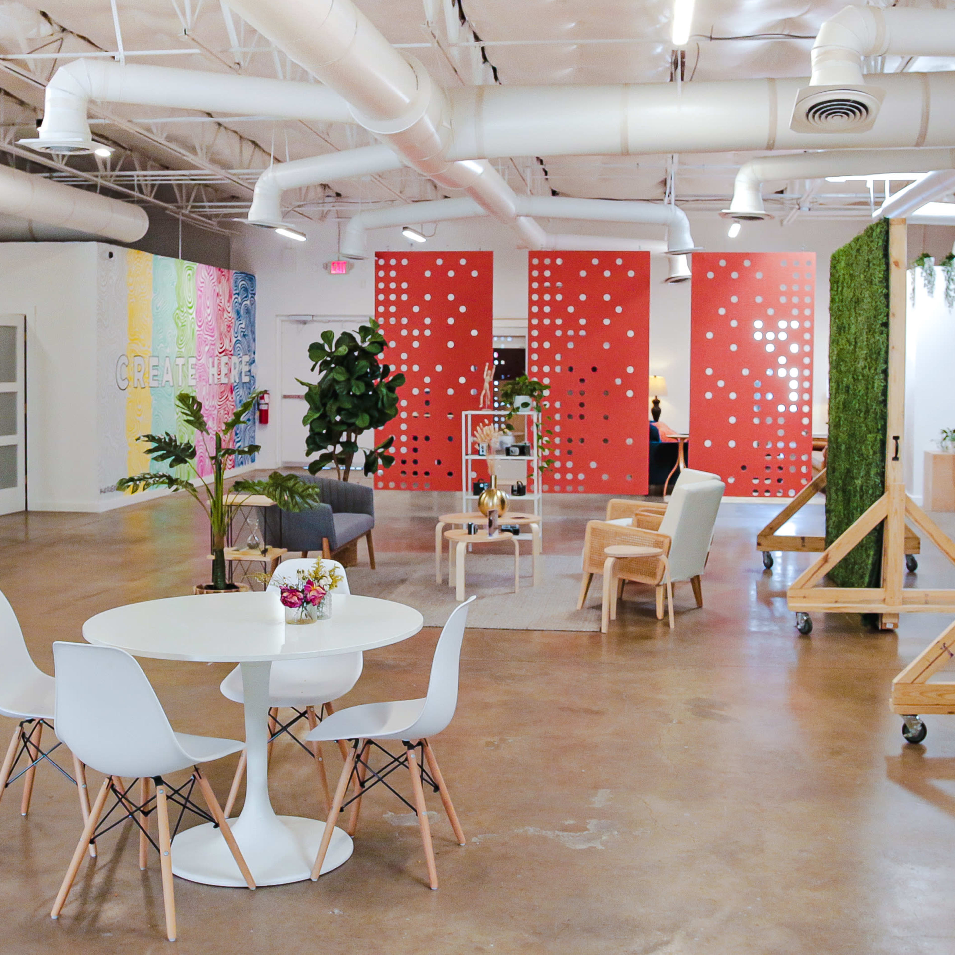 A modern workspace featuring a round table with white chairs, colorful accent walls, plants, and various seating areas.