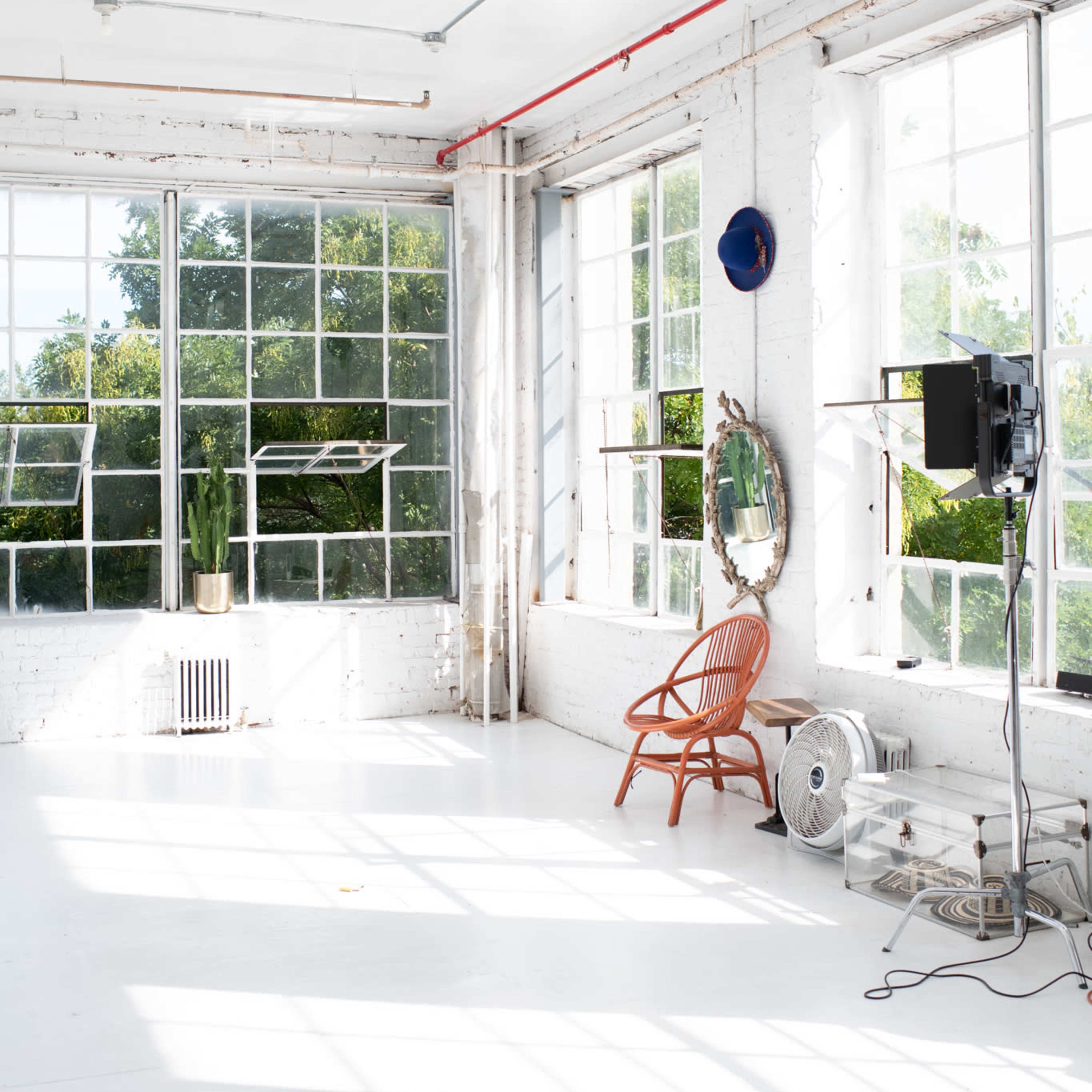 A sunlit studio features large windows, a simple rattan chair, a fan, and a mirror against a white wall.