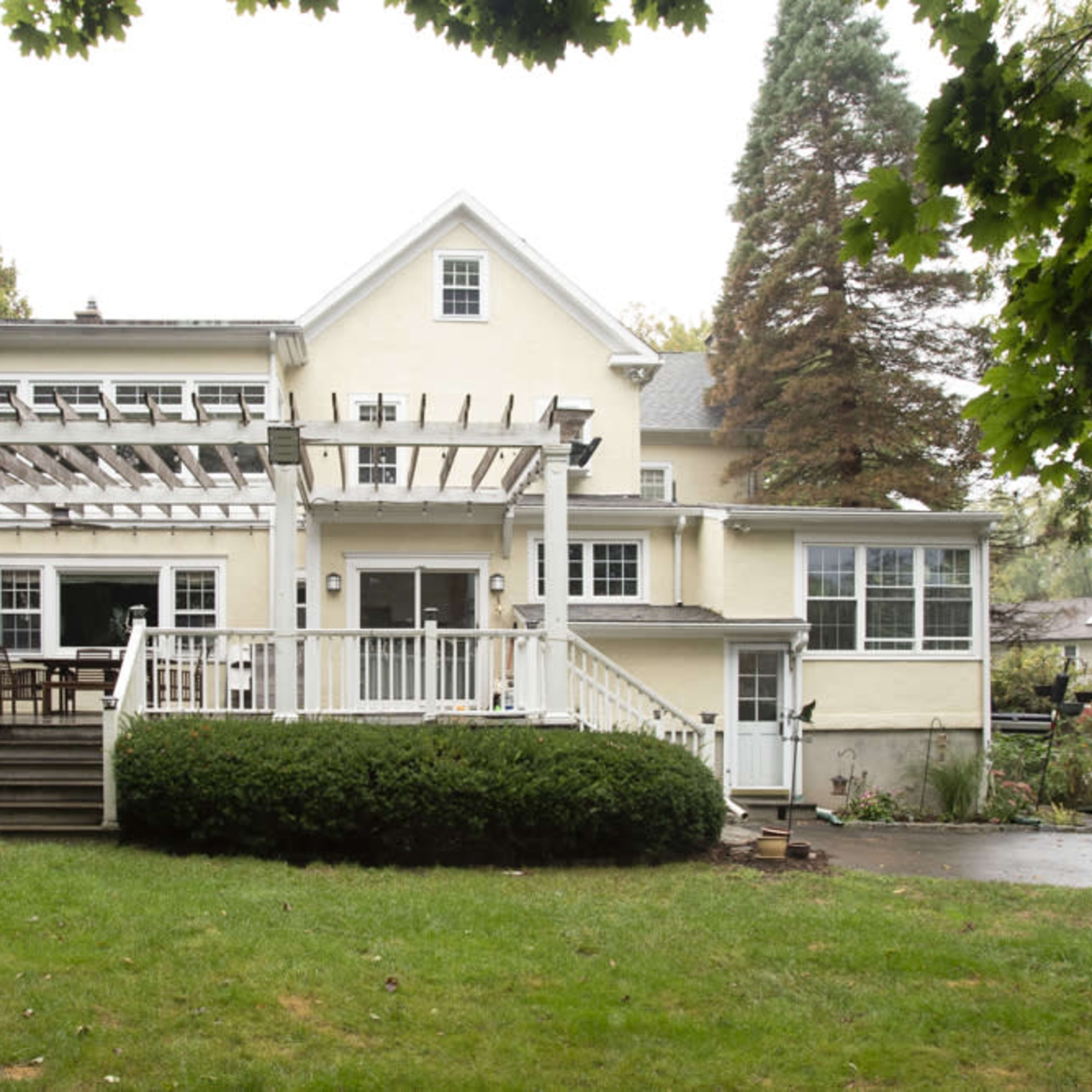 A two-story yellow house features a large wooden deck with a pergola, surrounded by green grass and shrubs.