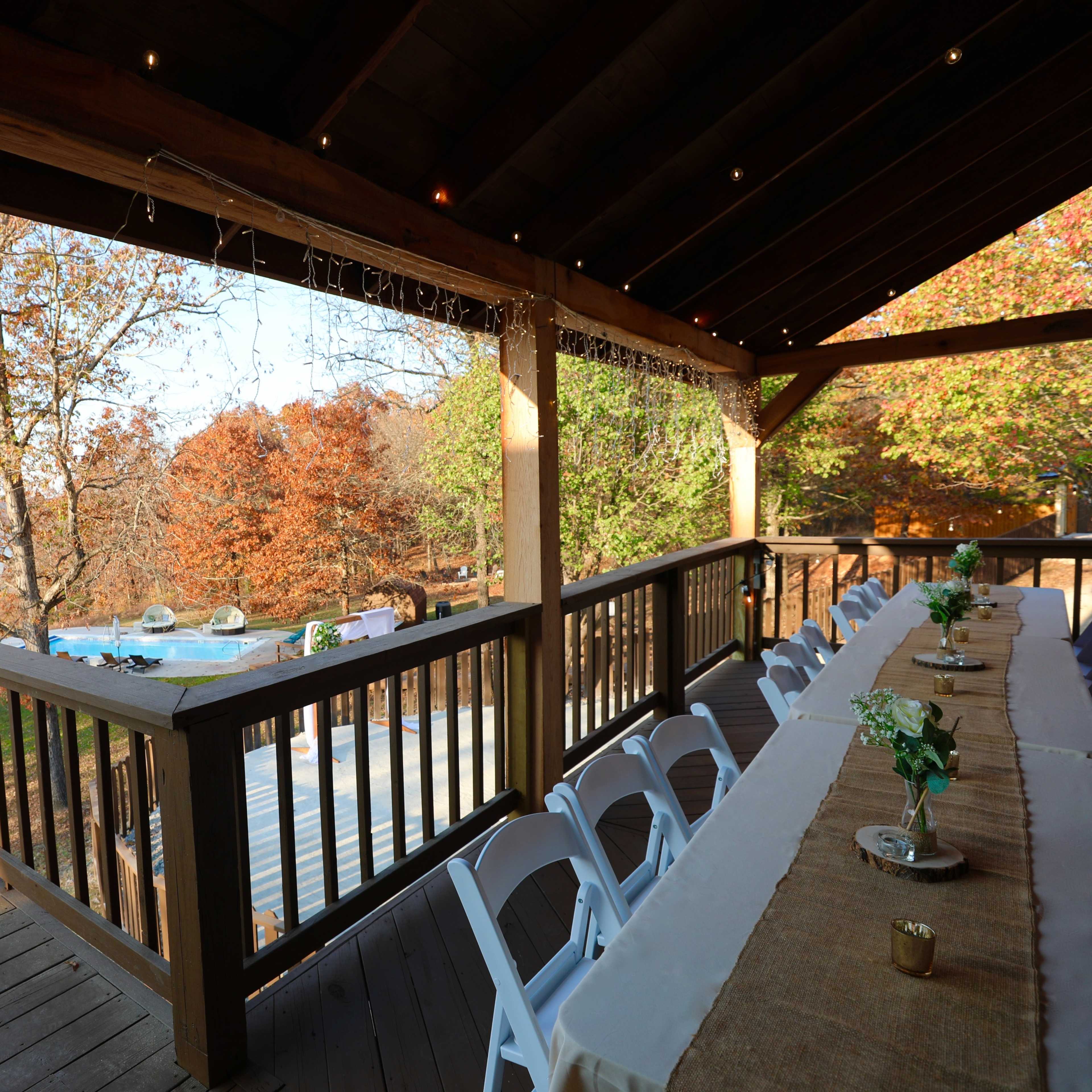 A wooden deck with a long table set for dining overlooks a scenic view of trees in autumn foliage and a body of water in the background.