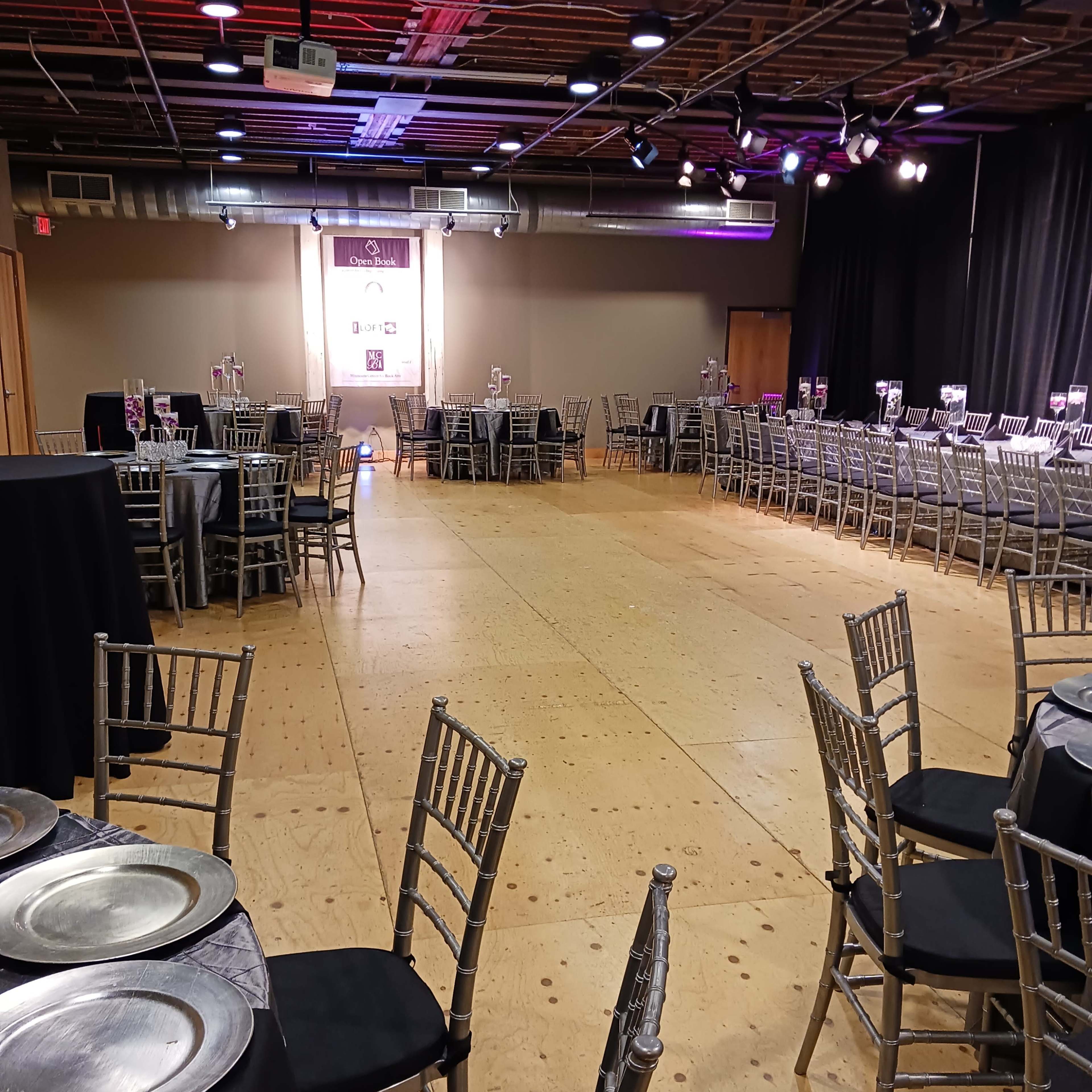 A spacious event hall arranged with tables and chairs, illuminated by overhead lights and a backdrop for a formal gathering.