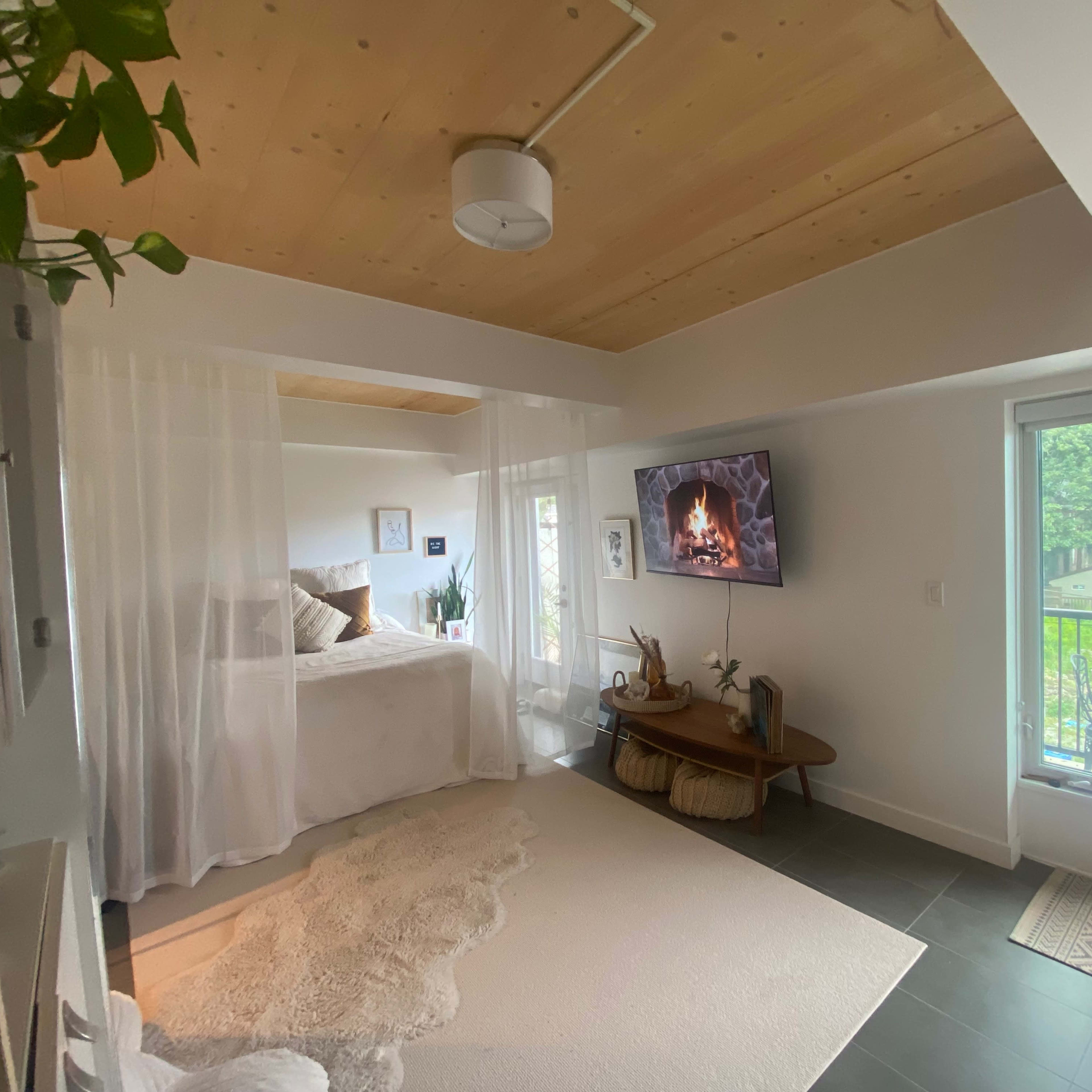 A cozy room features a bed draped with sheer curtains, a wall-mounted television displaying a fireplace, and a balcony door leading to an outdoor view.
