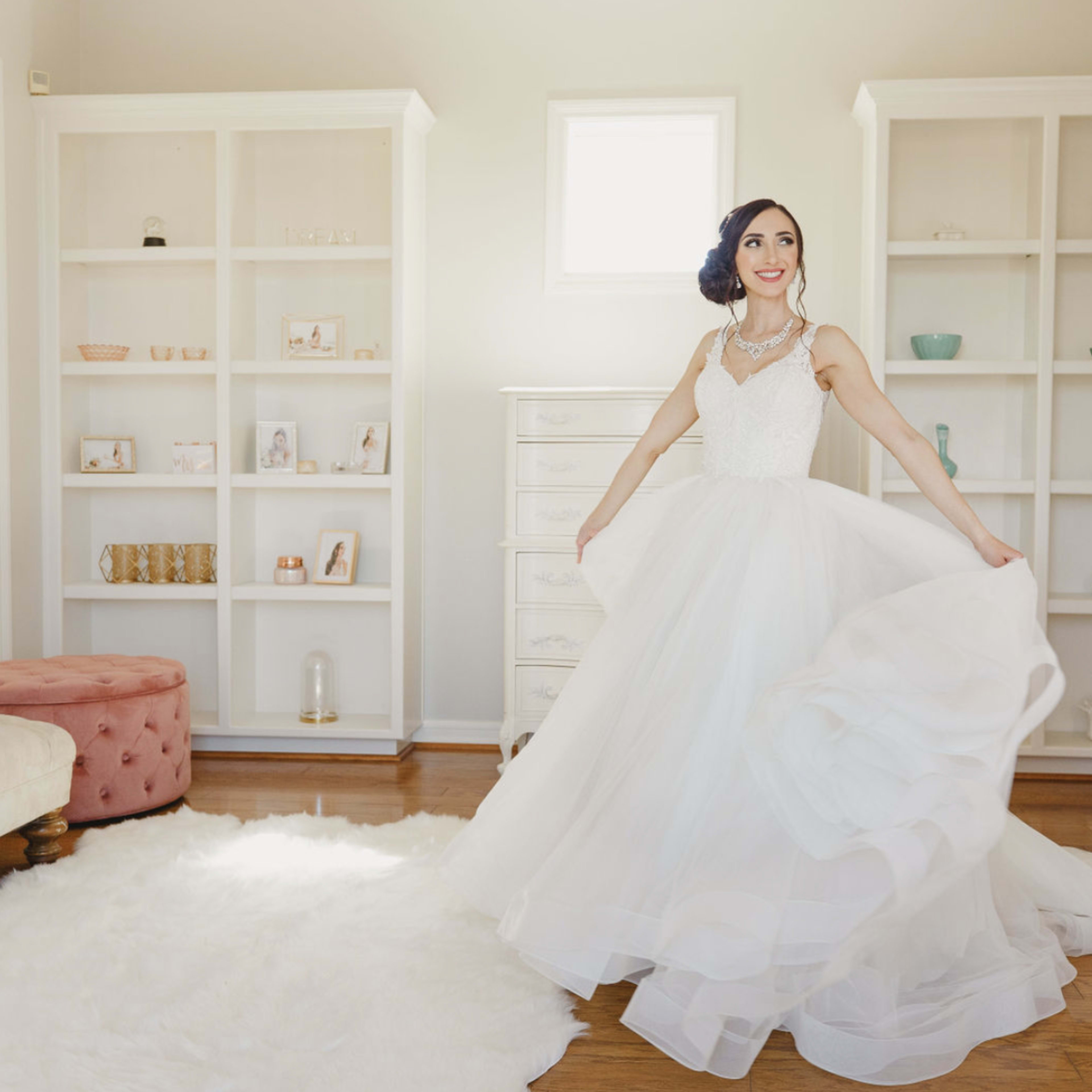 A bride wearing a white gown twirls in a bright room with shelves displaying various decorative items and a plush sofa.