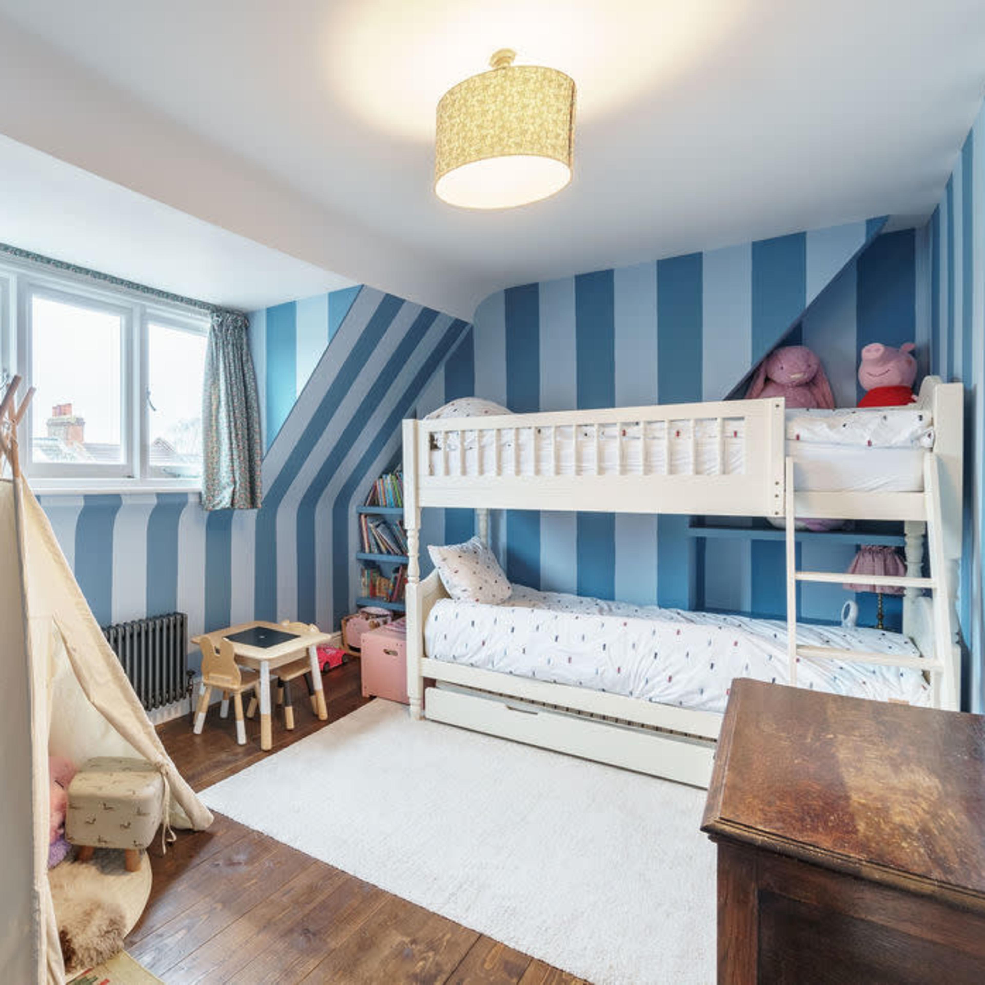 A children's bedroom features striped blue walls, a bunk bed, a play tent, and a cozy rug on a wooden floor.