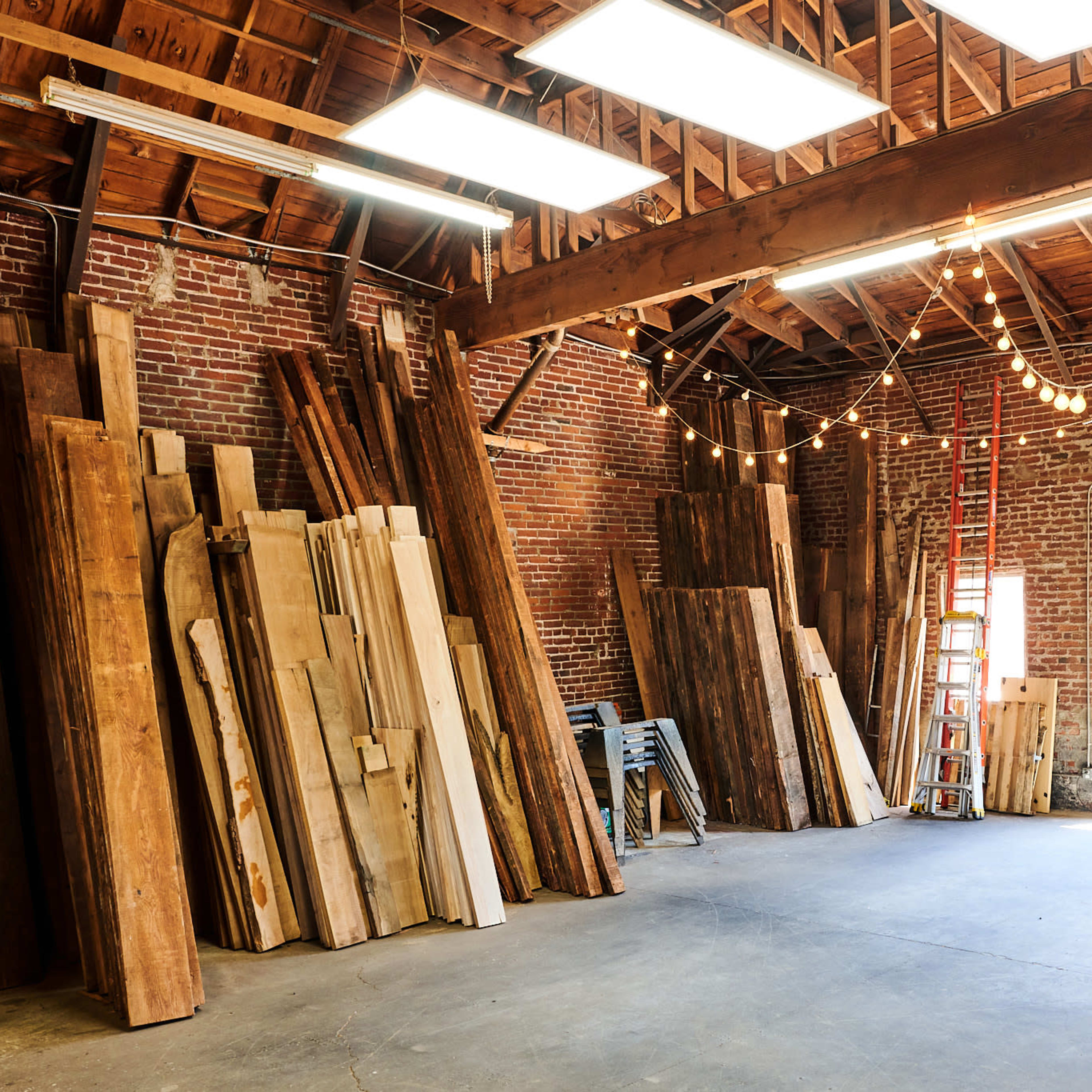 A spacious workshop with stacked wooden planks against the brick walls and overhead string lights illuminating the area.