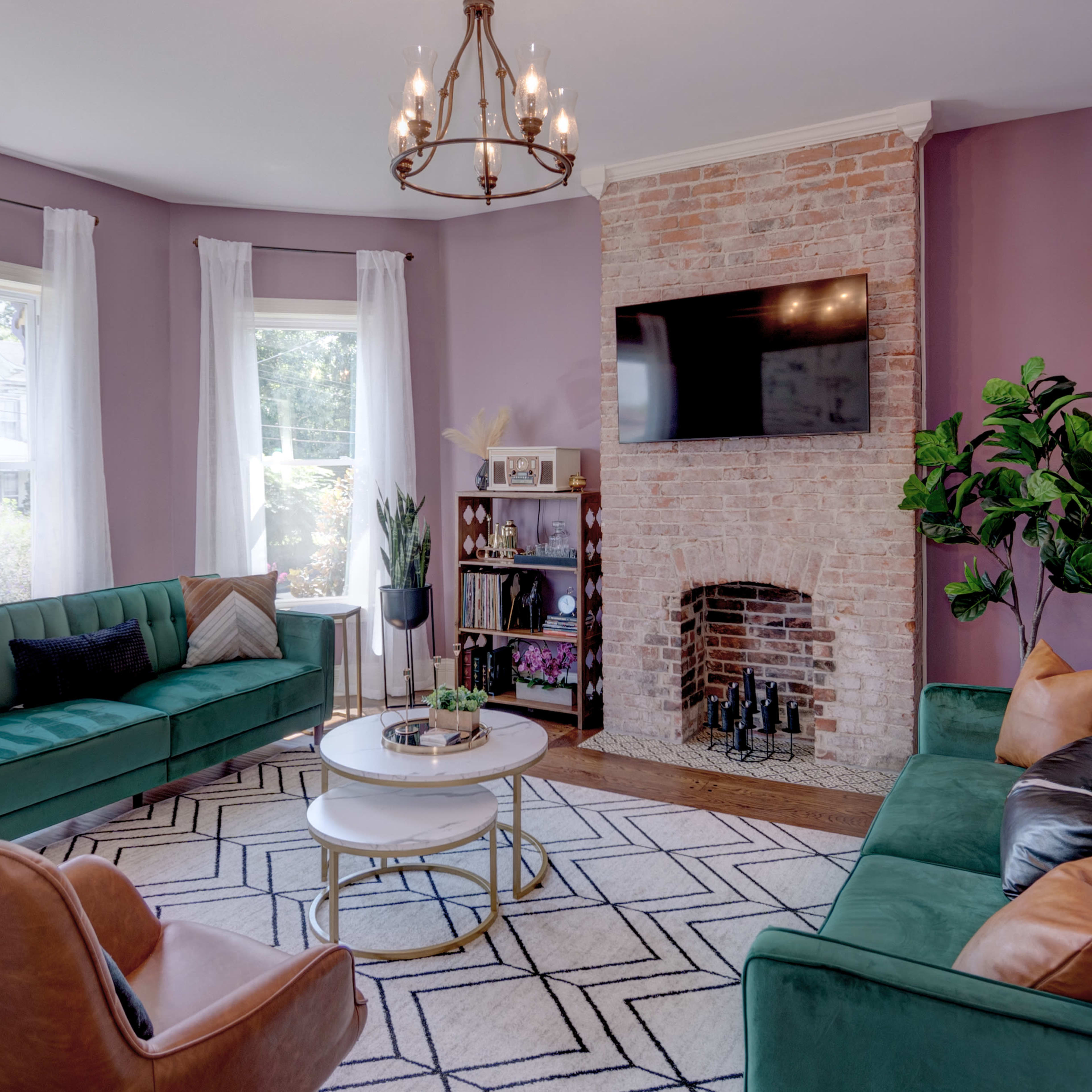 The living room features green sofas, a round coffee table, a brick fireplace, and large windows with white curtains.