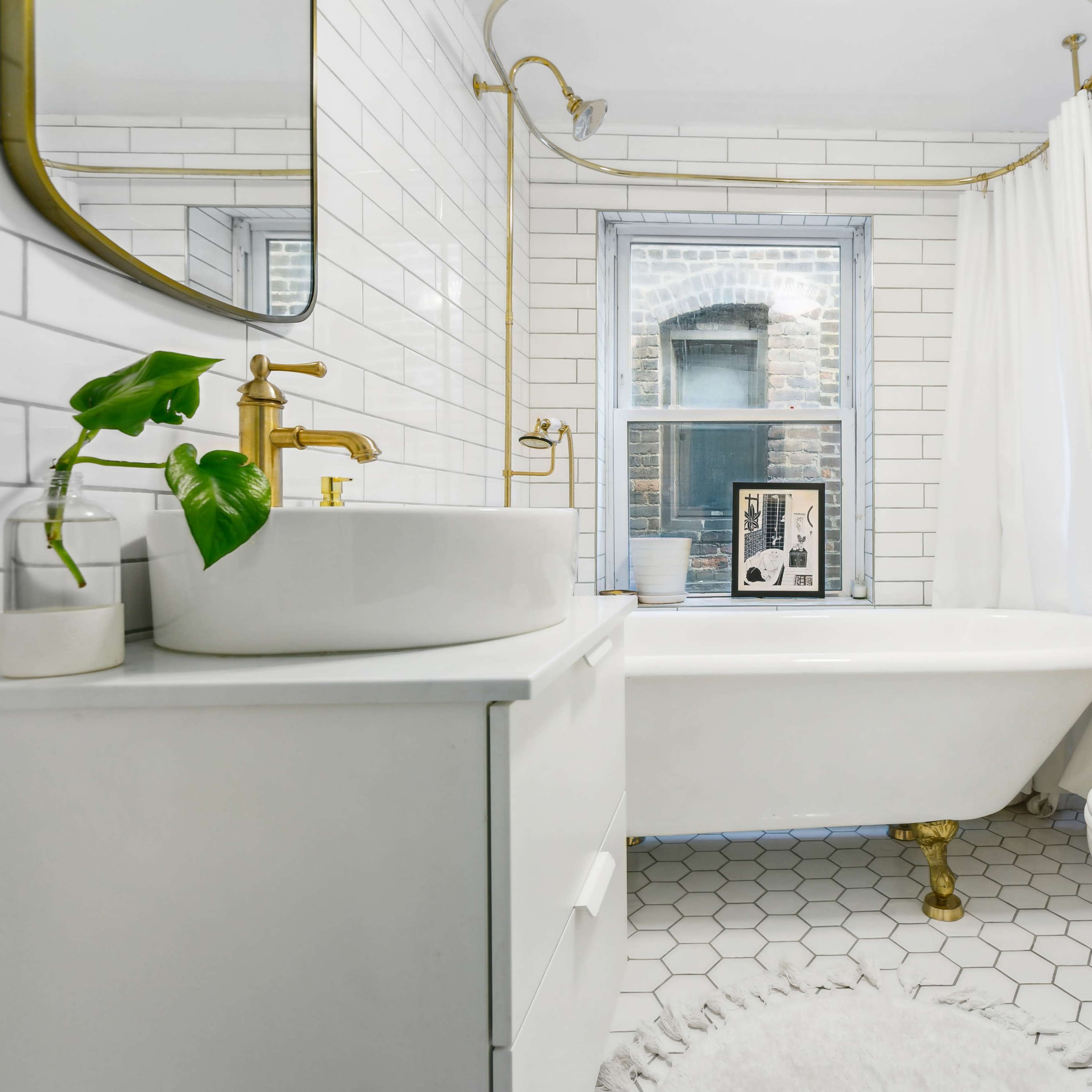 A modern bathroom features a freestanding tub, white tiled walls, a gold-accented mirror, and a large window allowing natural light.