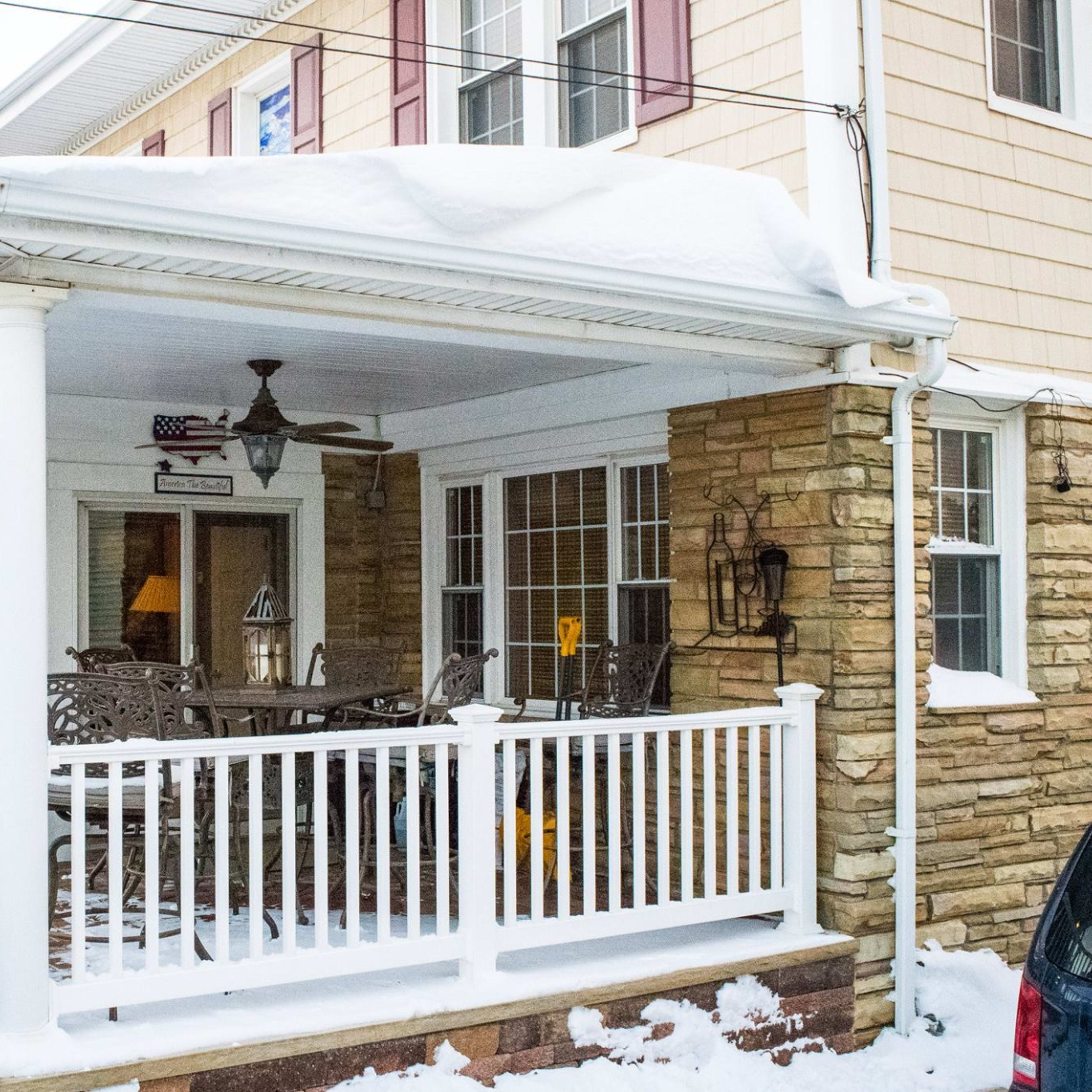 A snow-covered porch with white railings, a stone facade, and outdoor furniture, attached to a two-story house.