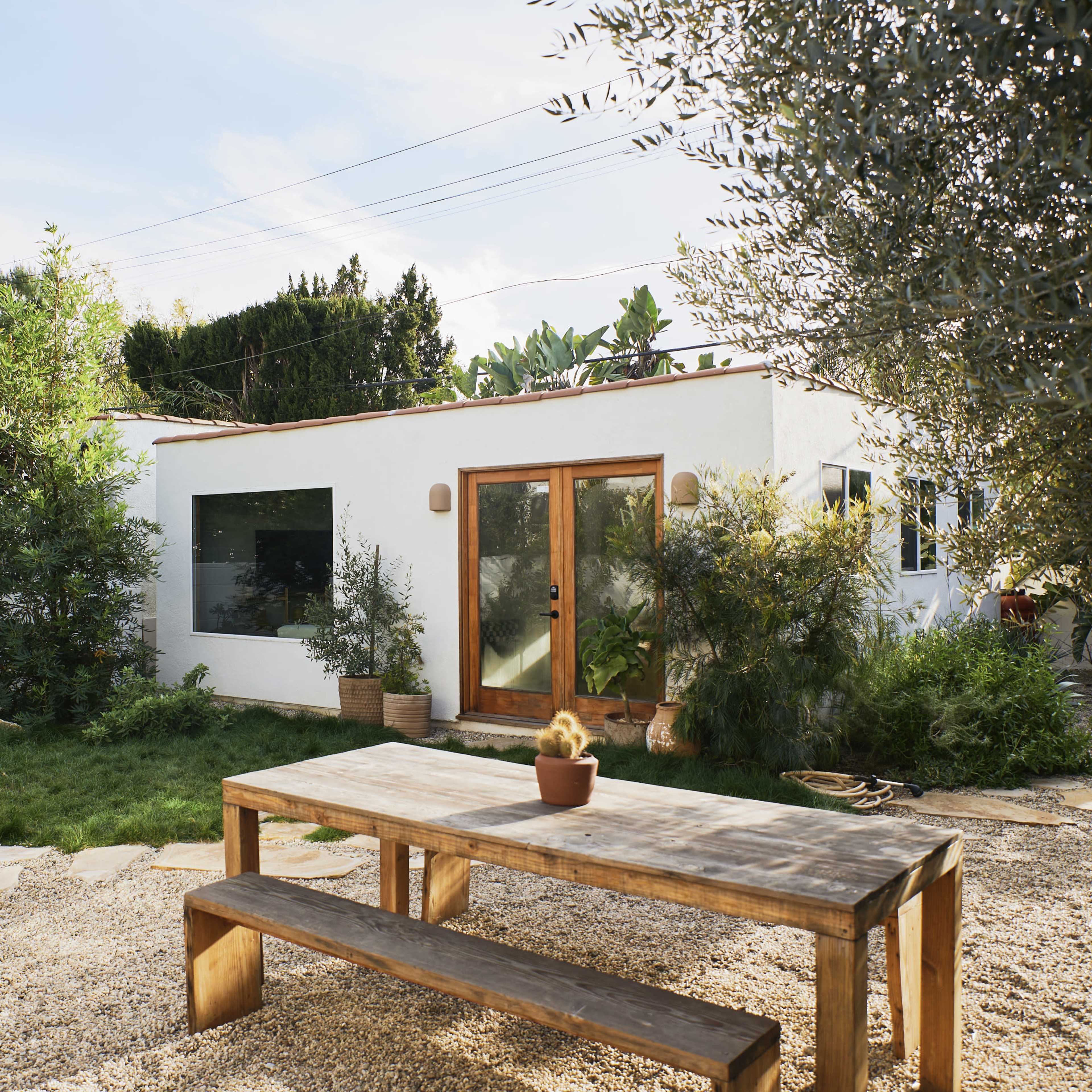 A modern, white house with large windows is surrounded by greenery and a gravel yard featuring a wooden picnic table.