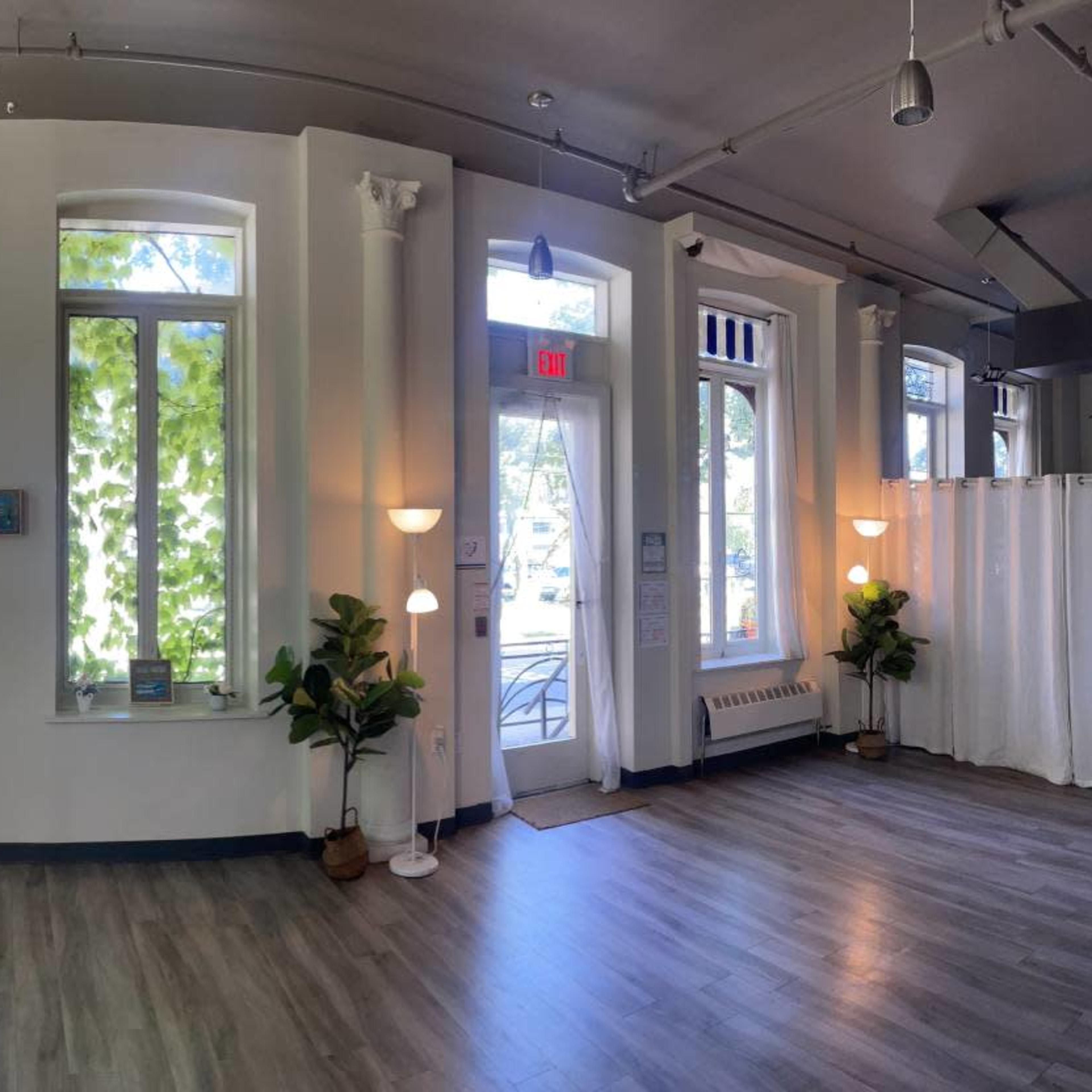 A spacious interior with large windows letting in natural light, potted plants in each corner, and an entrance door leading outside.
