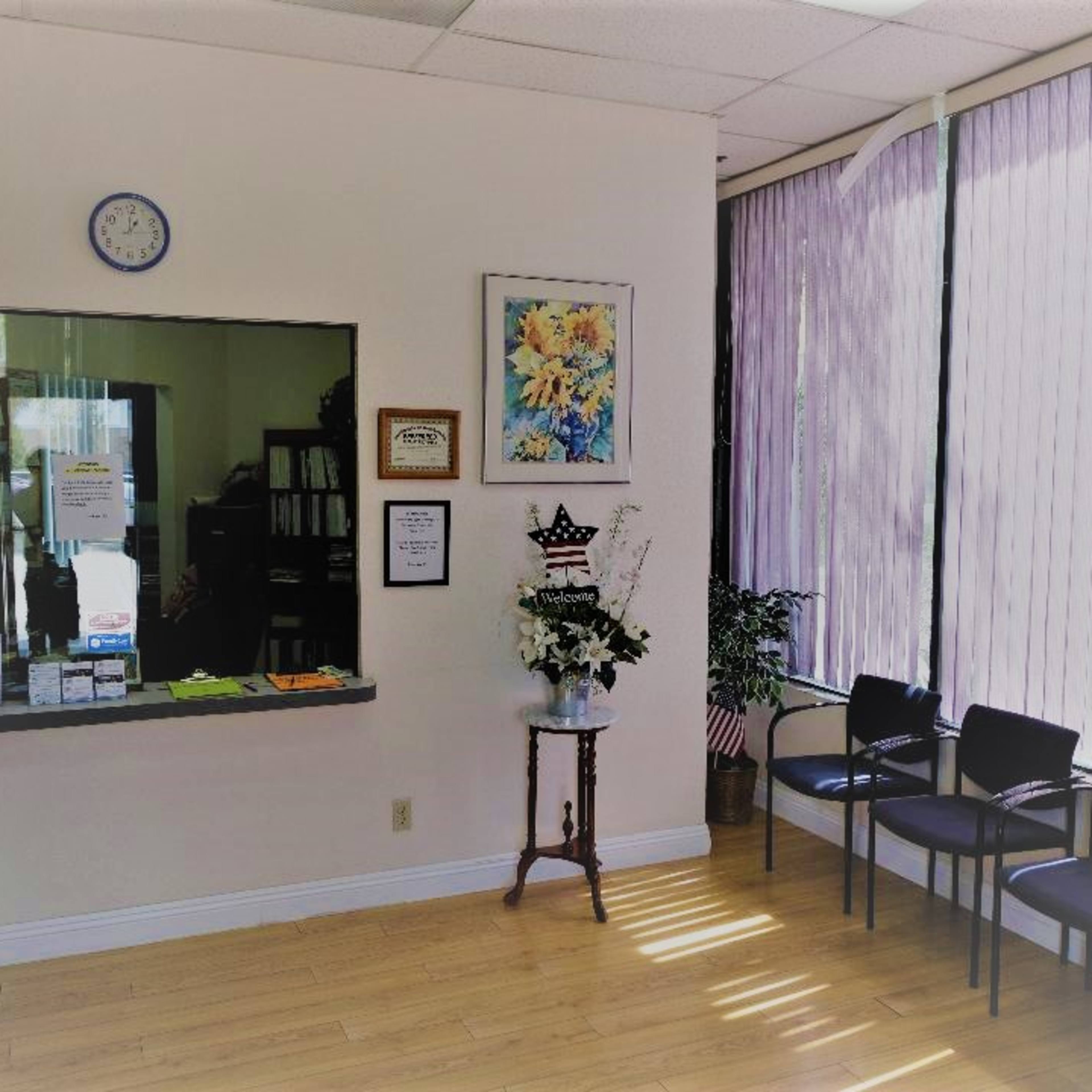 A waiting room with a counter, a floral display, and several black chairs arranged in front of large windows with purple curtains.