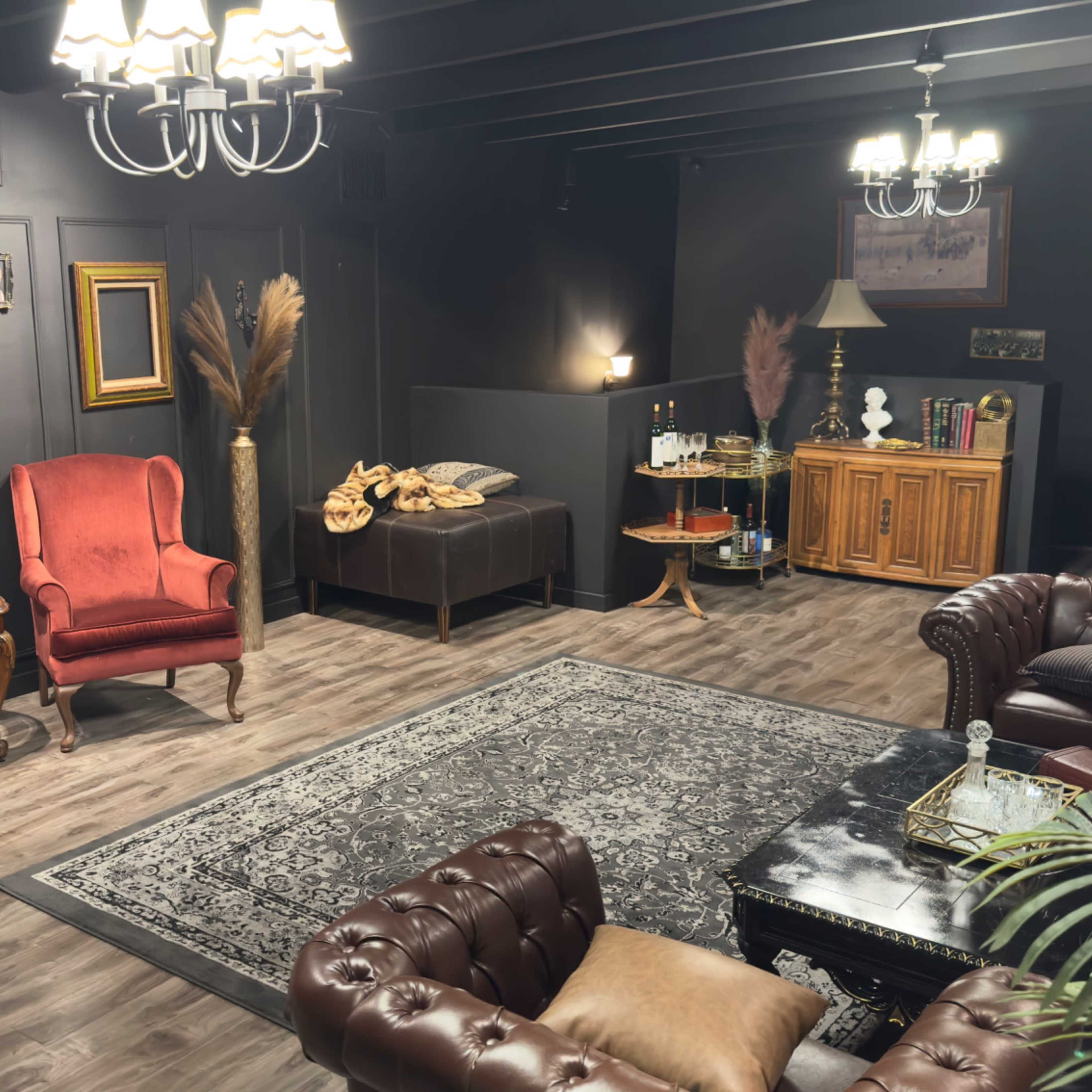 The scene features a cozy, well-decorated room with two red armchairs, a dark sofa, a wooden cabinet, and a decorative rug, illuminated by chandeliers.