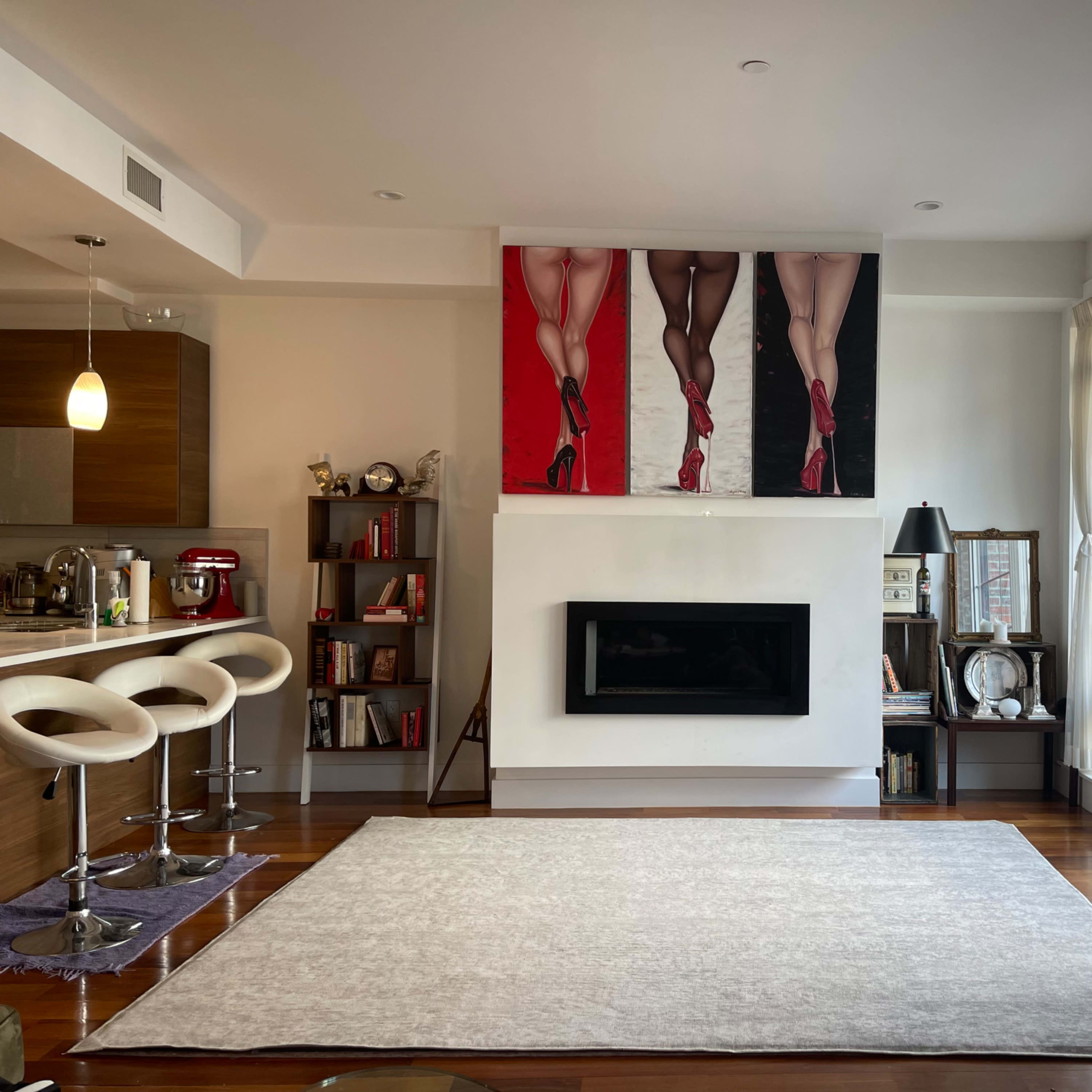 A modern living room features a large artwork of legs in high heels above a sleek fireplace, surrounded by warm wood flooring and contemporary furniture.