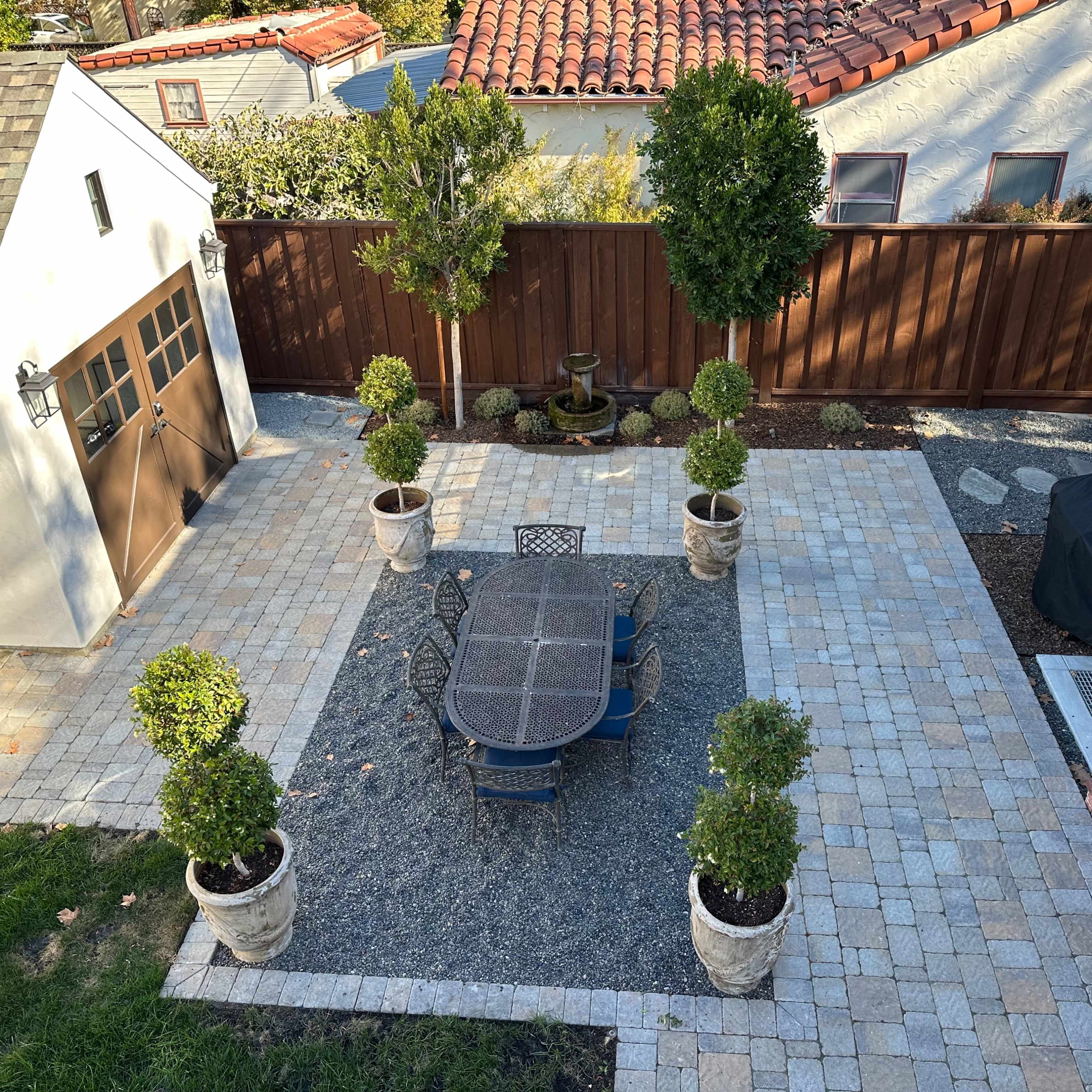 A landscaped patio area featuring a large stone table surrounded by chairs, flanked by potted plants and bordered by a wooden fence.