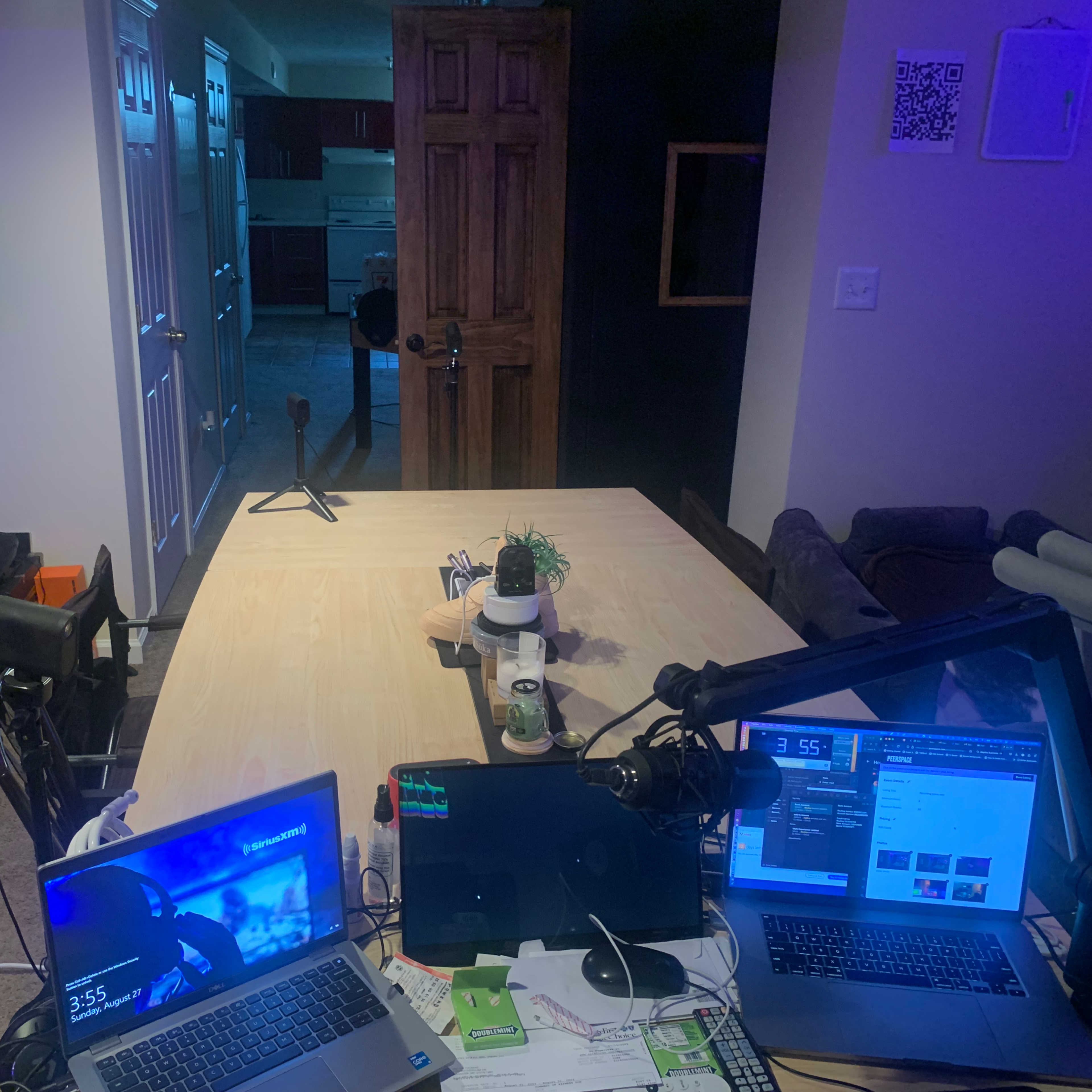 A workspace is set up on a table with three laptops, a microphone stand, and a small plant, alongside a visible kitchen area in the background.