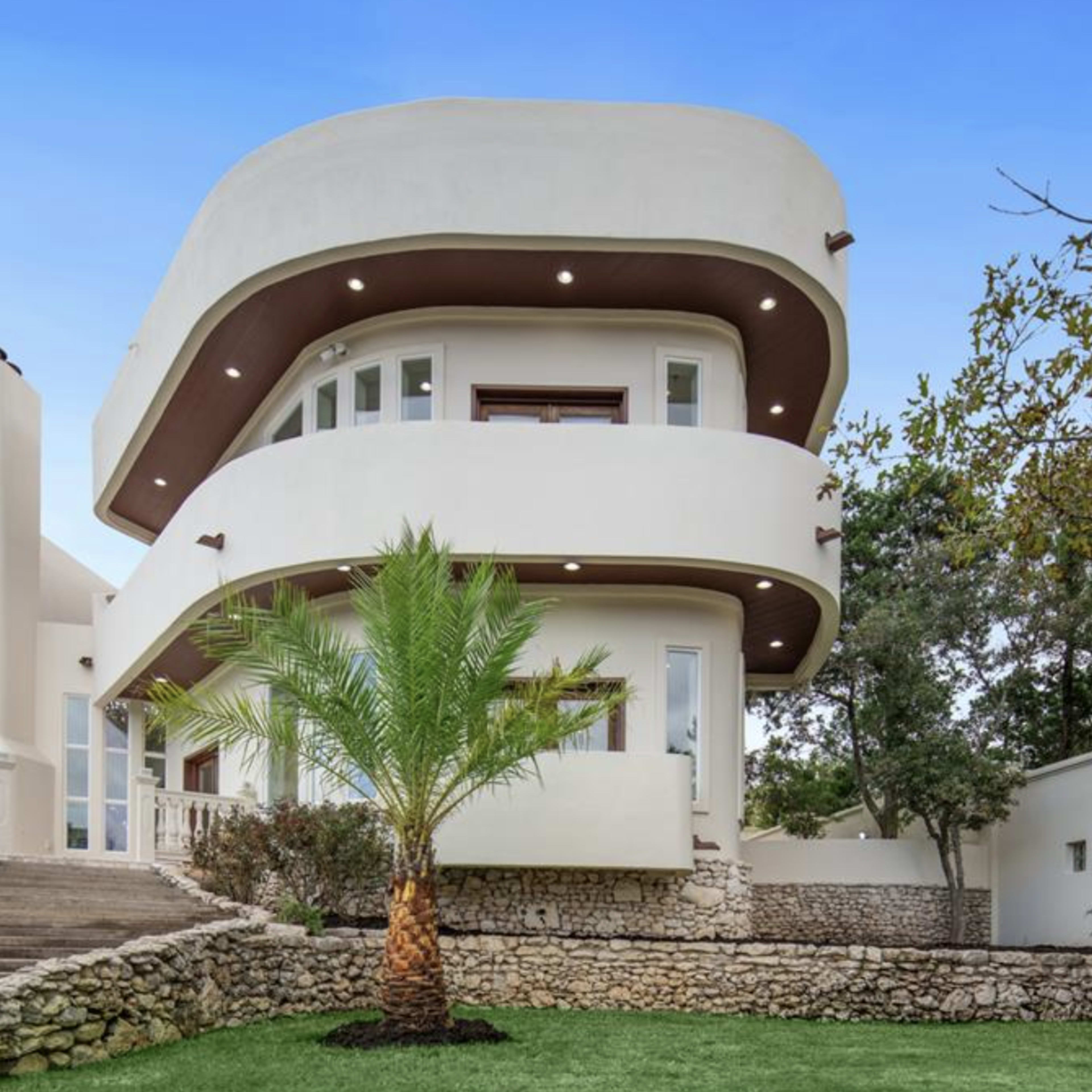 A modern two-story house with a curved architectural design, surrounded by a landscaped yard featuring a palm tree and stone retaining walls.