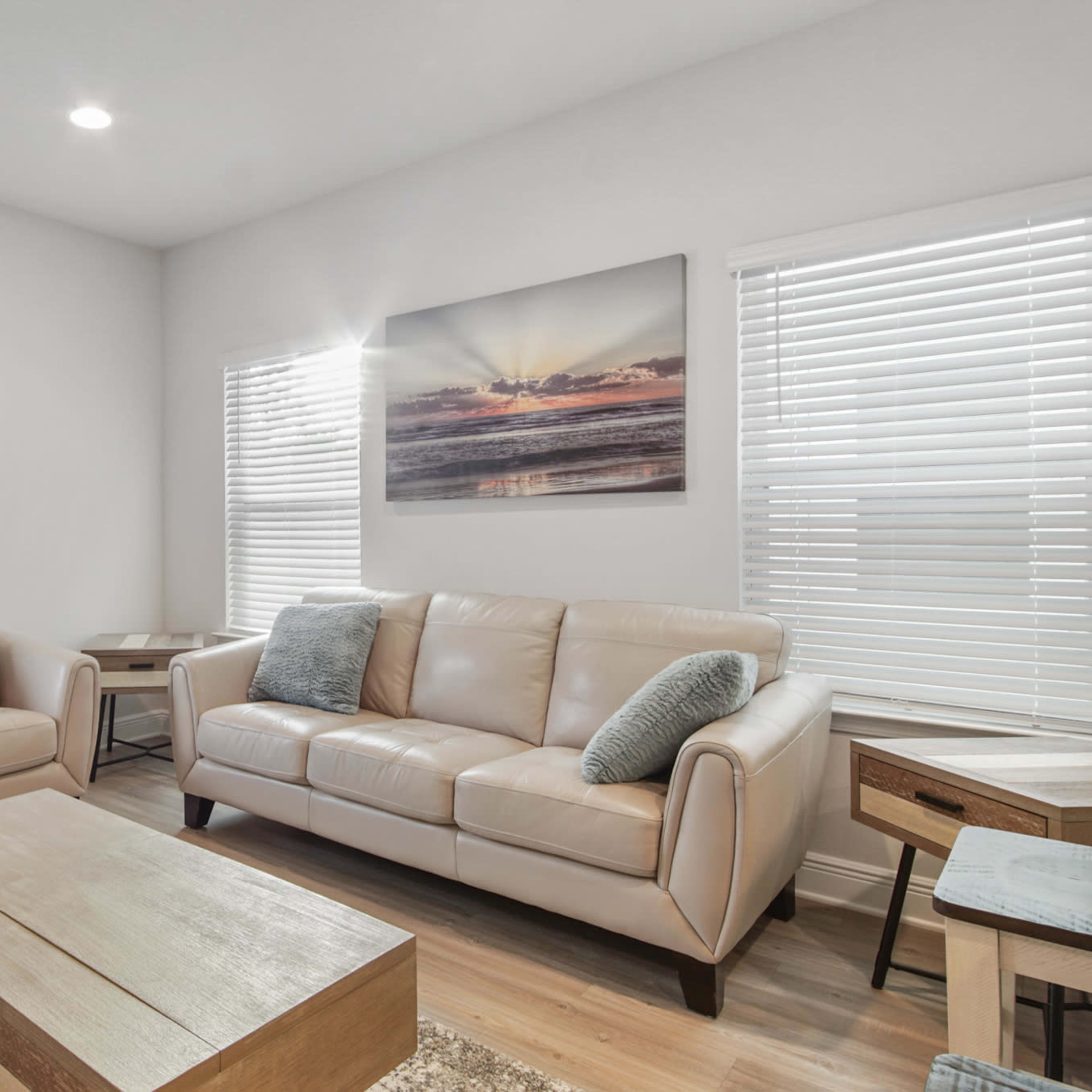 A bright, modern living room featuring a light-colored leather sofa, a wooden coffee table, and large windows with blinds.