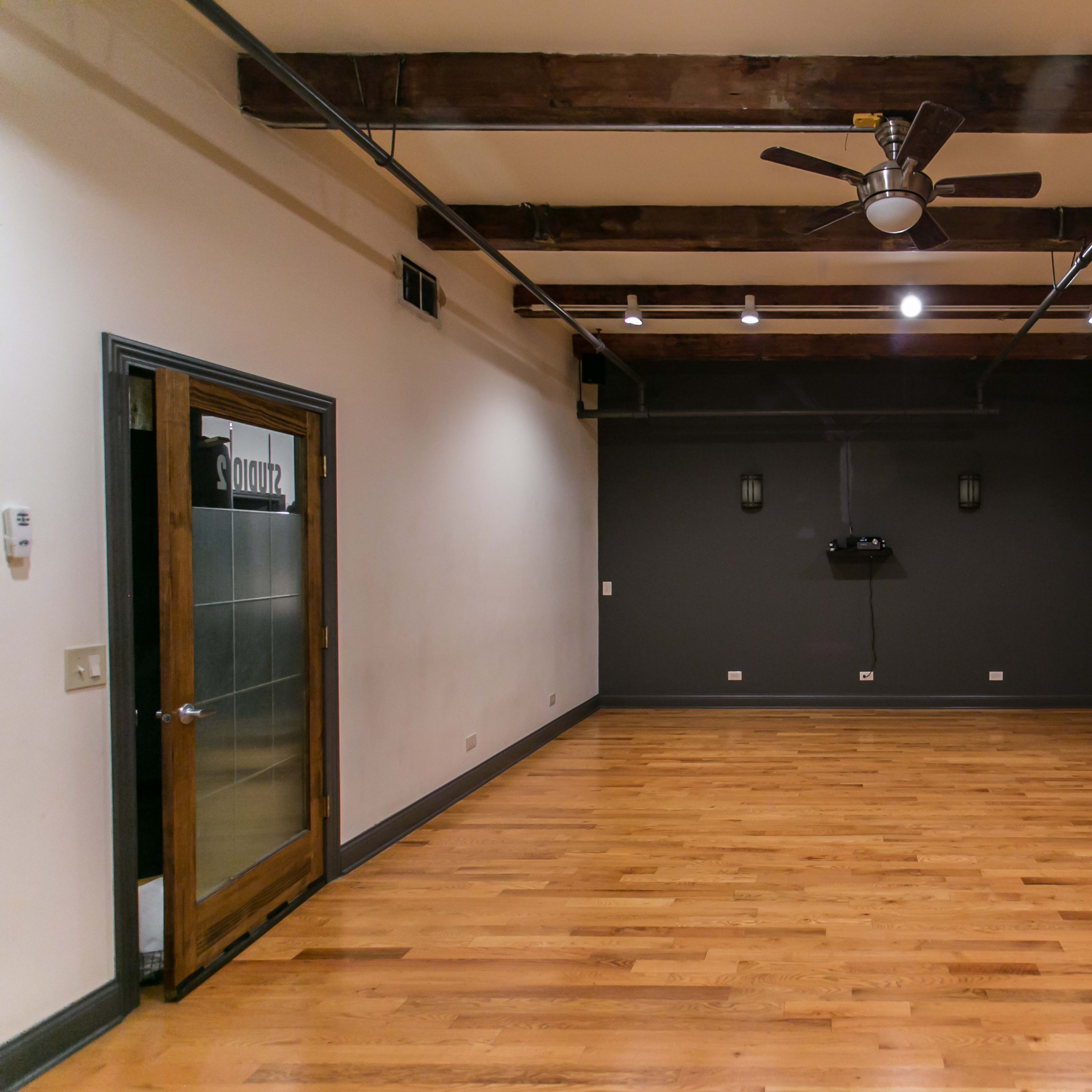 A spacious room with hardwood floors, exposed brick walls, and wooden beams, featuring a ceiling fan and a door with a glass panel.