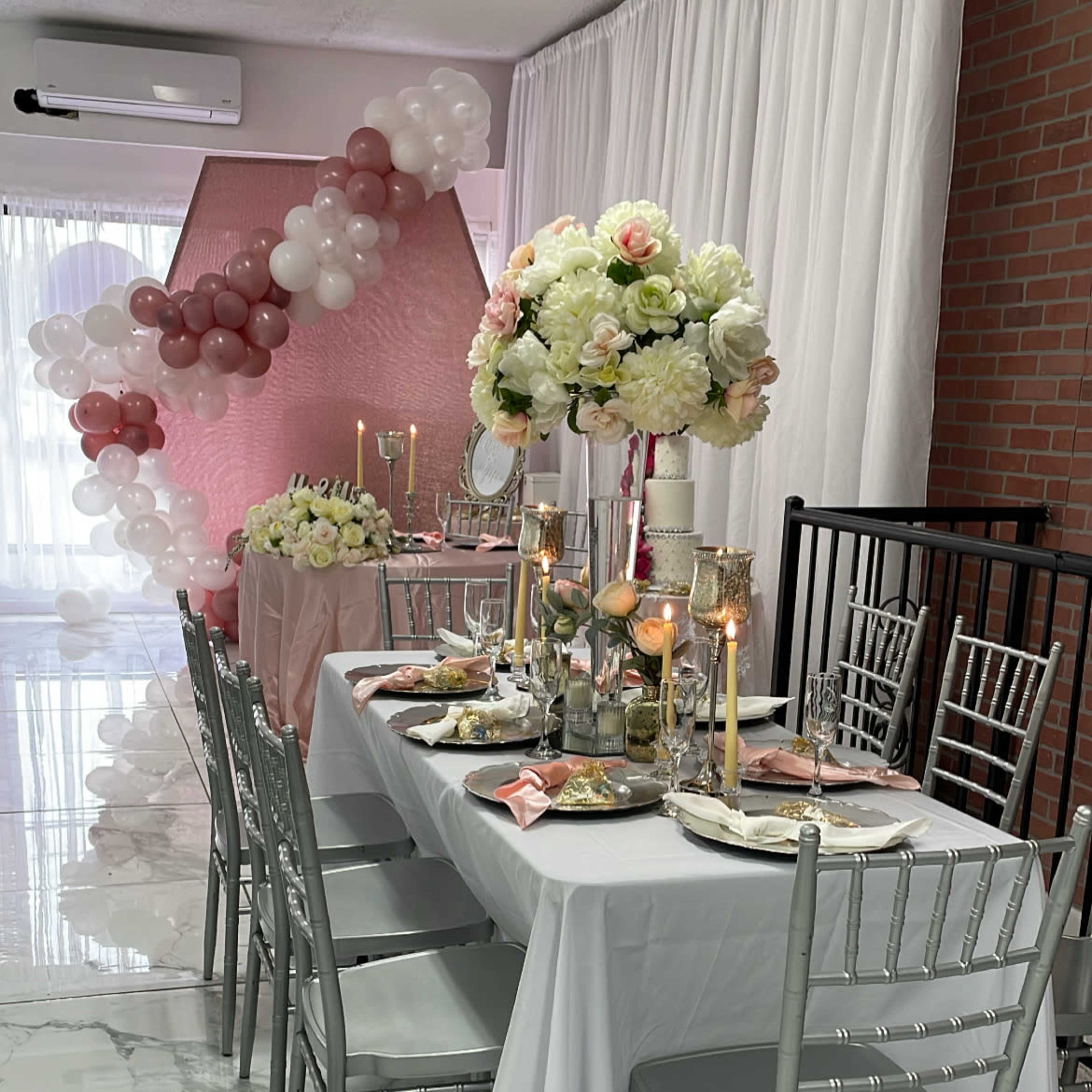 A decorated dining area features a long white table with elegant chairs, floral centerpieces, and candles, set against a backdrop of pink and white balloons.