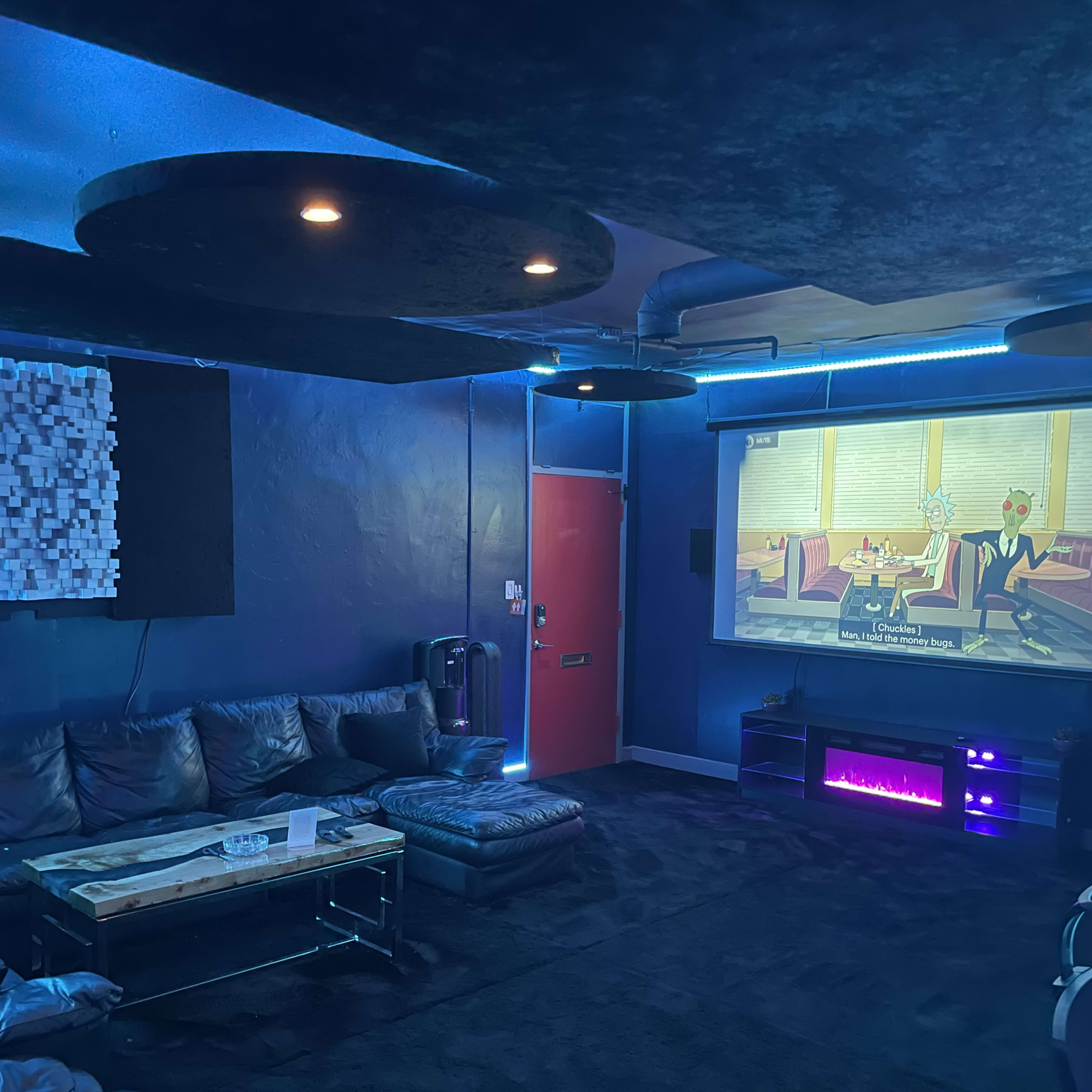 A dimly lit entertainment room featuring a large projection screen displaying a cartoon, with a black sofa set and a modern, illuminated entertainment unit.