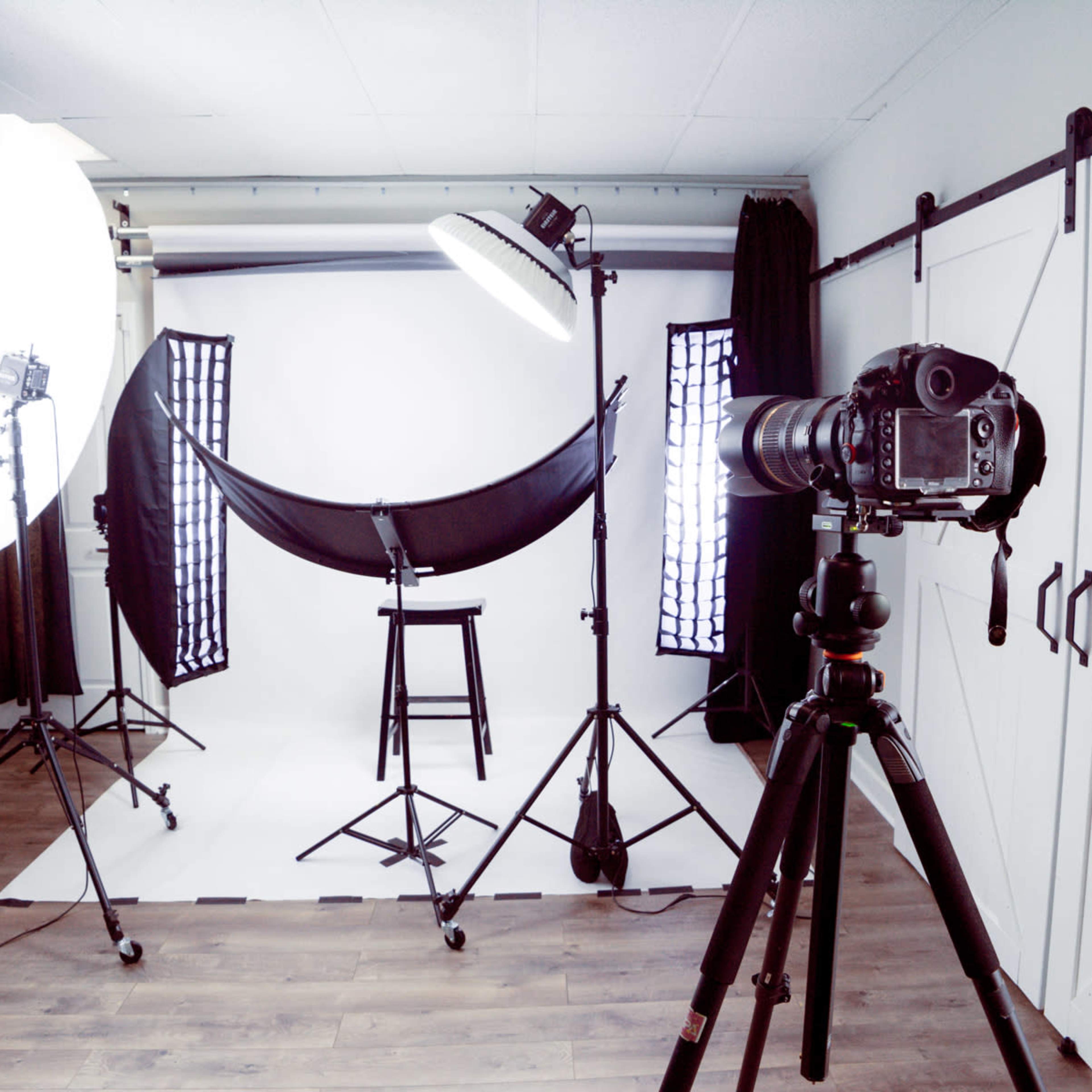 A photography studio setup with large softbox lights, a backdrop on stands, a stool, and a camera mounted on a tripod.