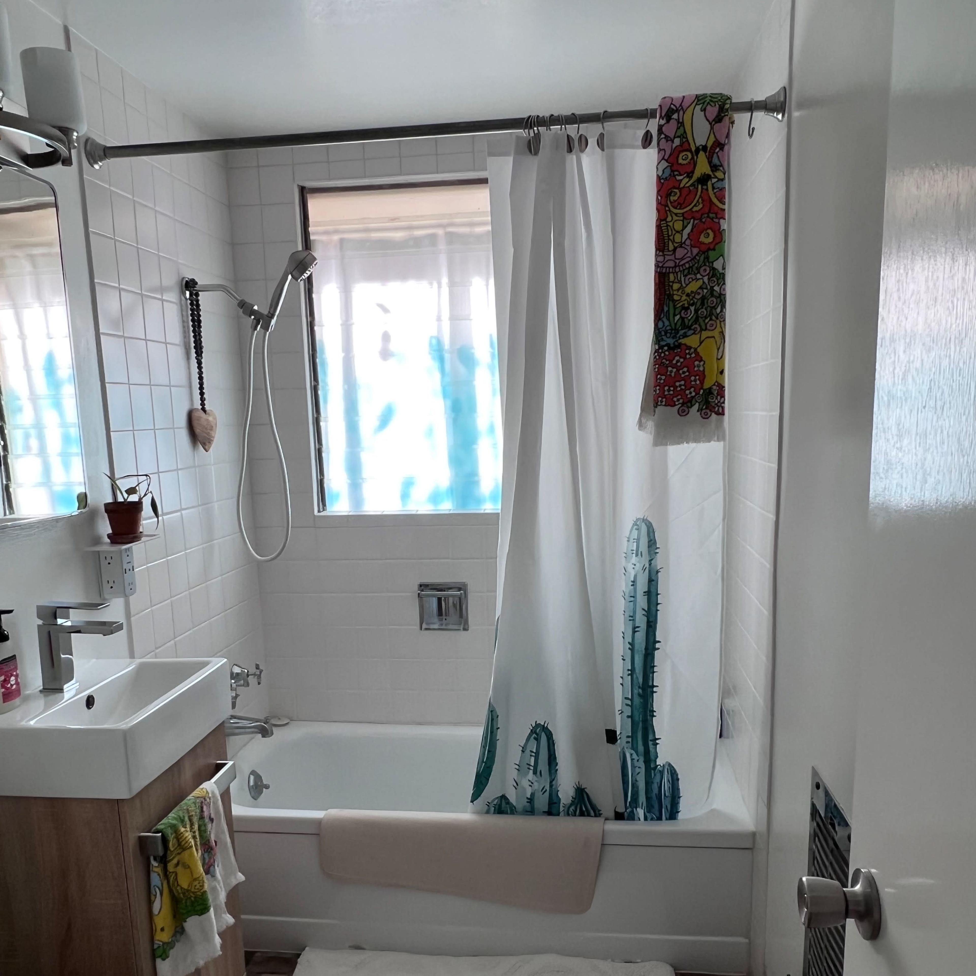 A bathroom with a bathtub, a shower curtain featuring cacti designs, a sink with a wooden cabinet, and a small rug on the floor.