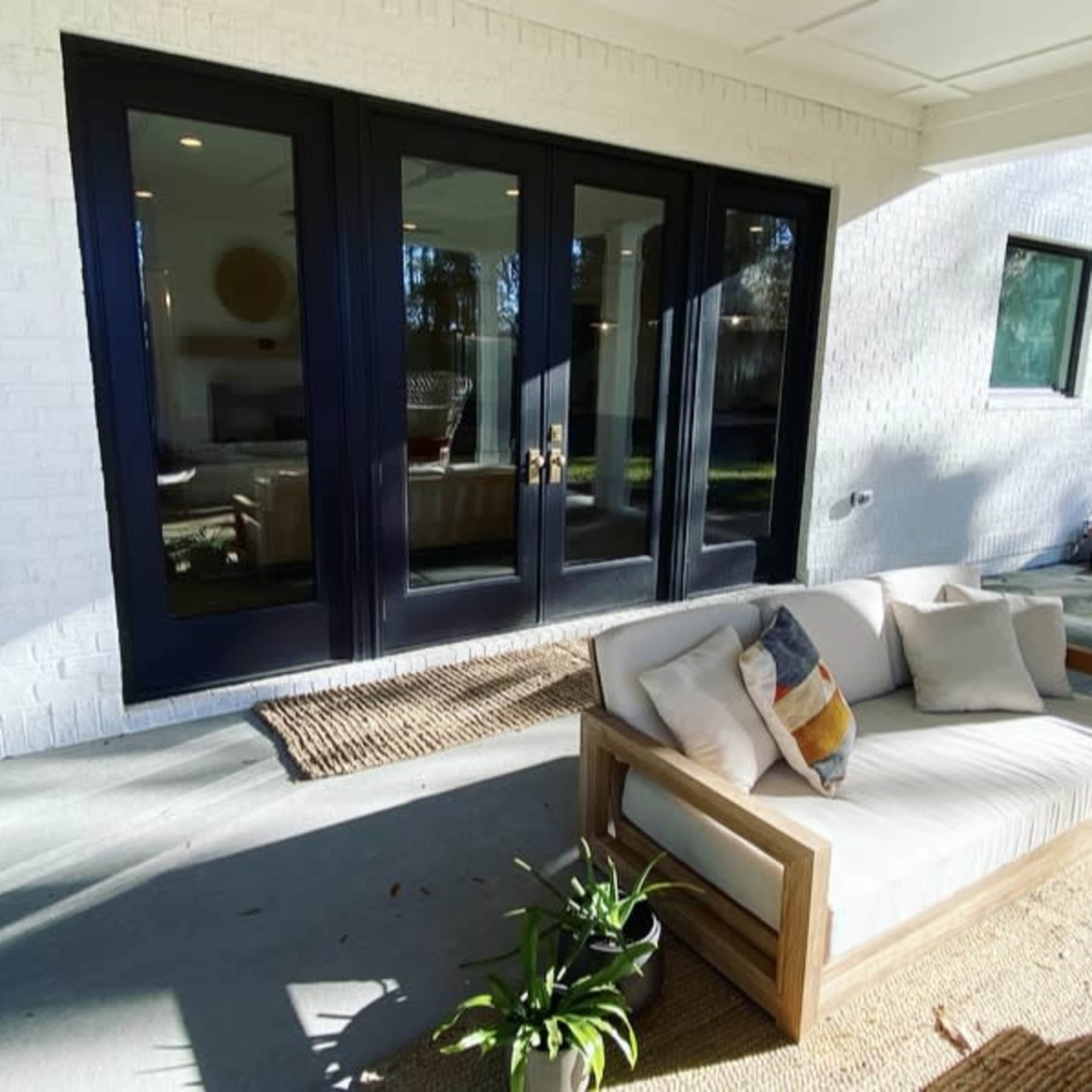 A wooden sofa with cushions sits on a concrete patio in front of large black-framed glass doors that open to a backyard.