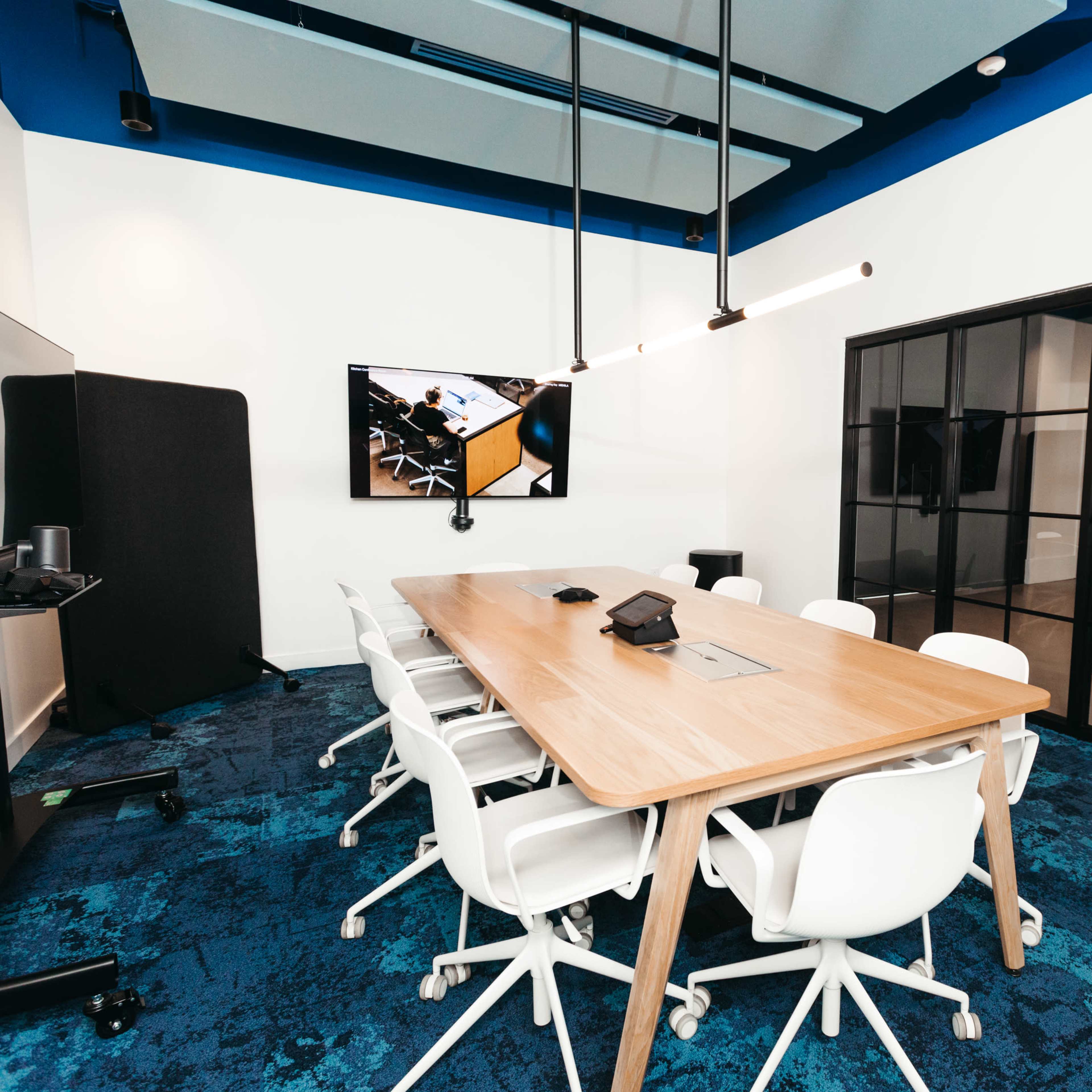 A modern conference room features a long wooden table surrounded by white chairs, with a large screen mounted on the wall and glass paneling.
