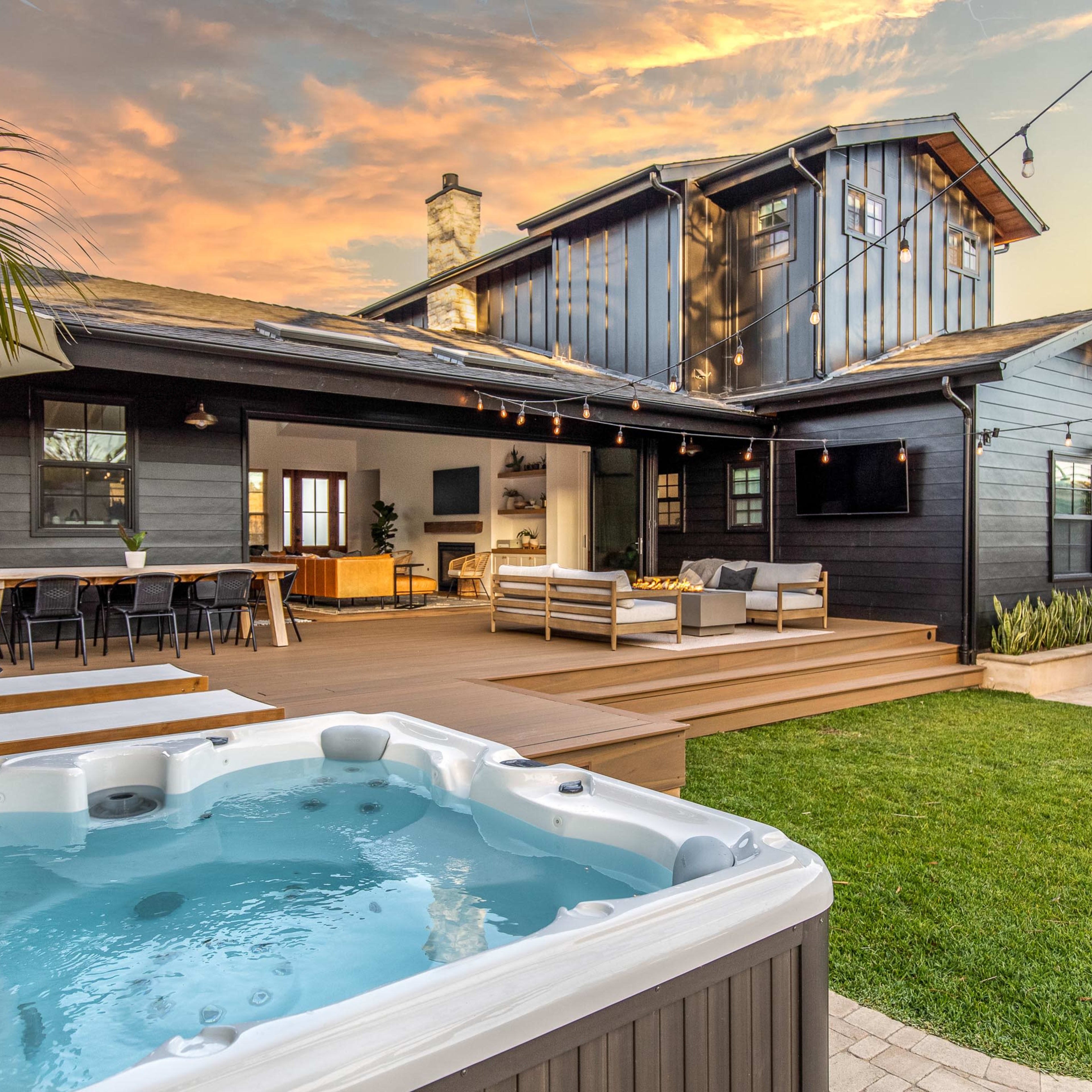 A modern outdoor space with a hot tub, wooden deck, dining area, and a contemporary home against a sunset sky.