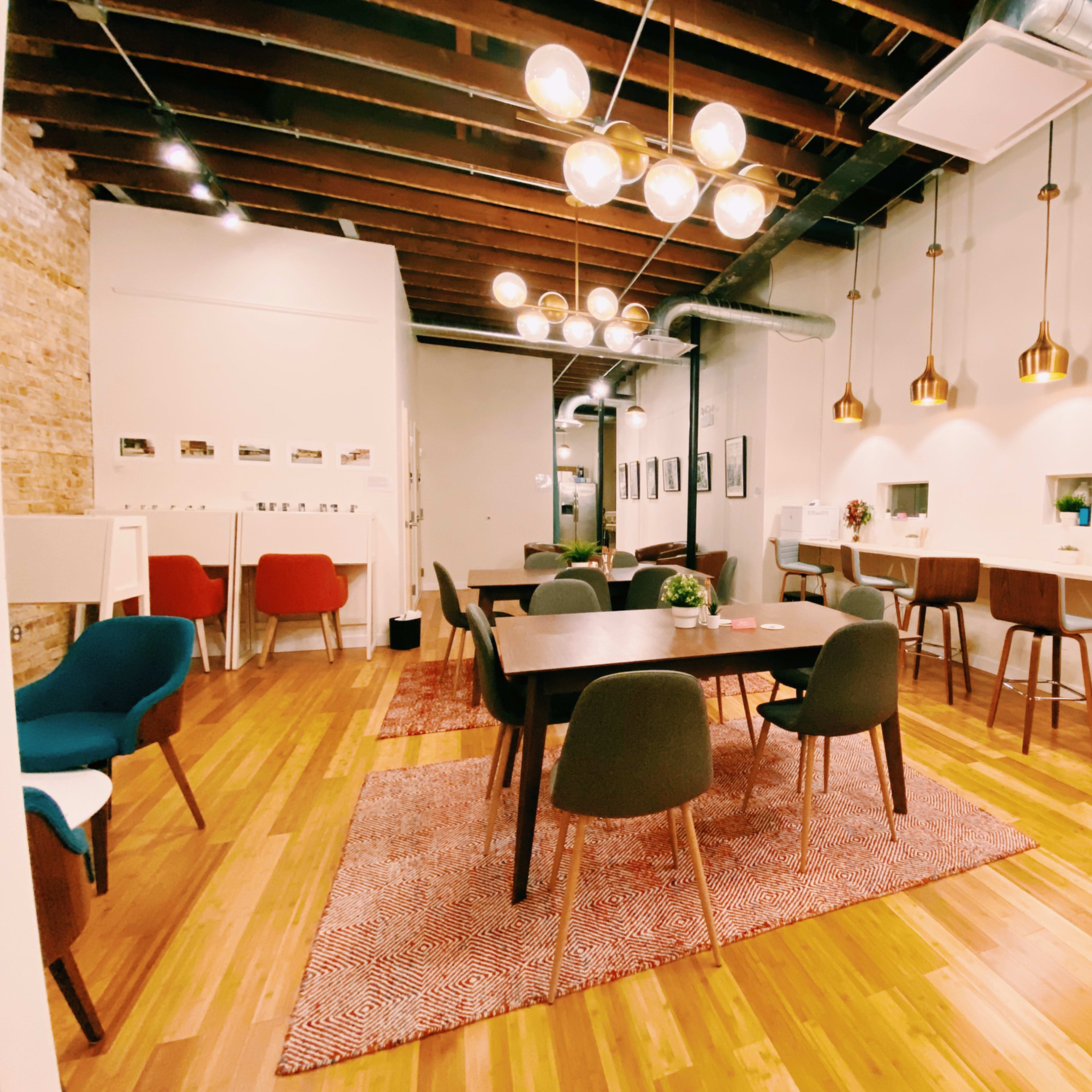 A modern, open workspace featuring a mix of tables, chairs, and a cozy seating area, complemented by decorative lighting and wooden flooring.