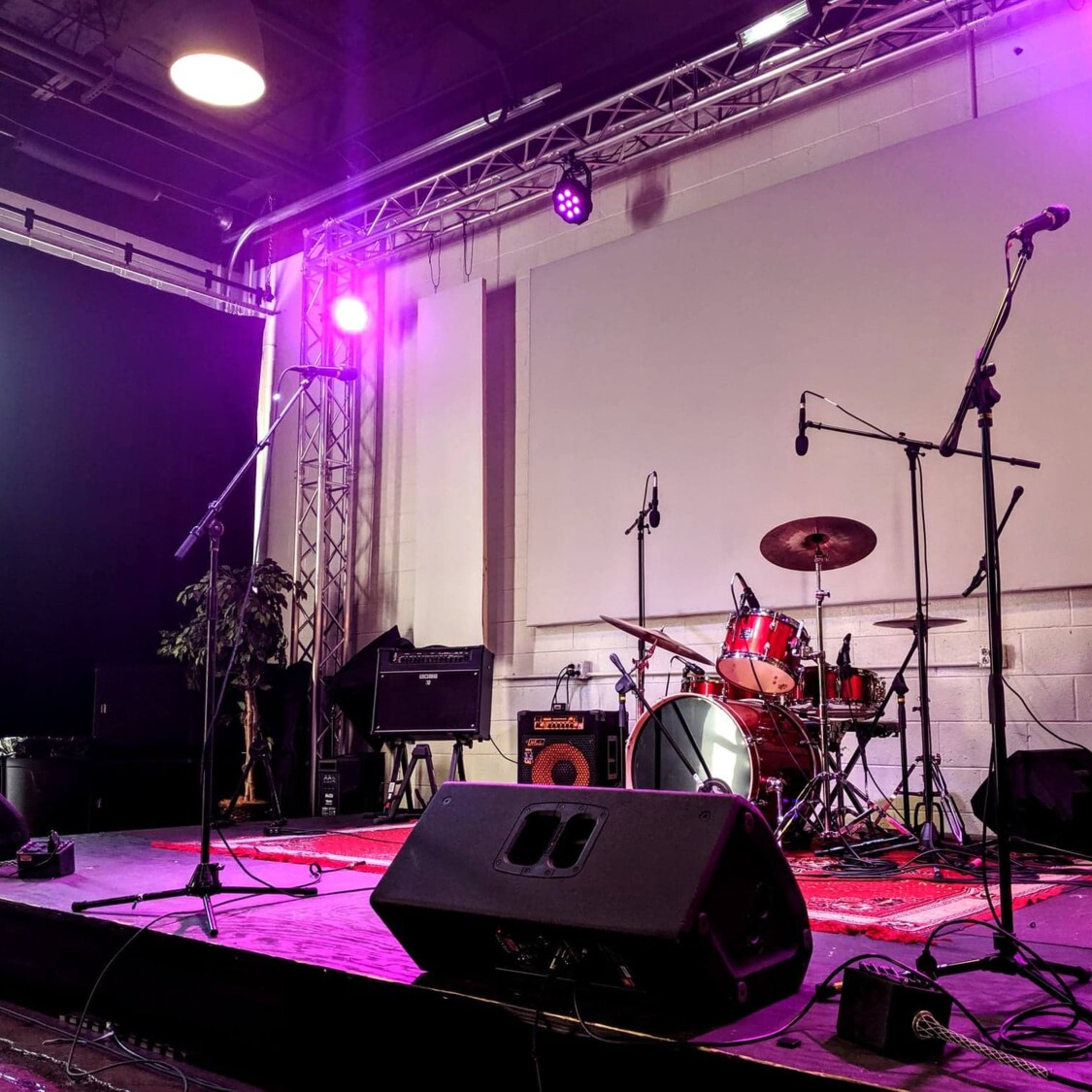 A stage is set up for a performance, featuring a drum kit, microphones, and amplifiers under purple lighting.