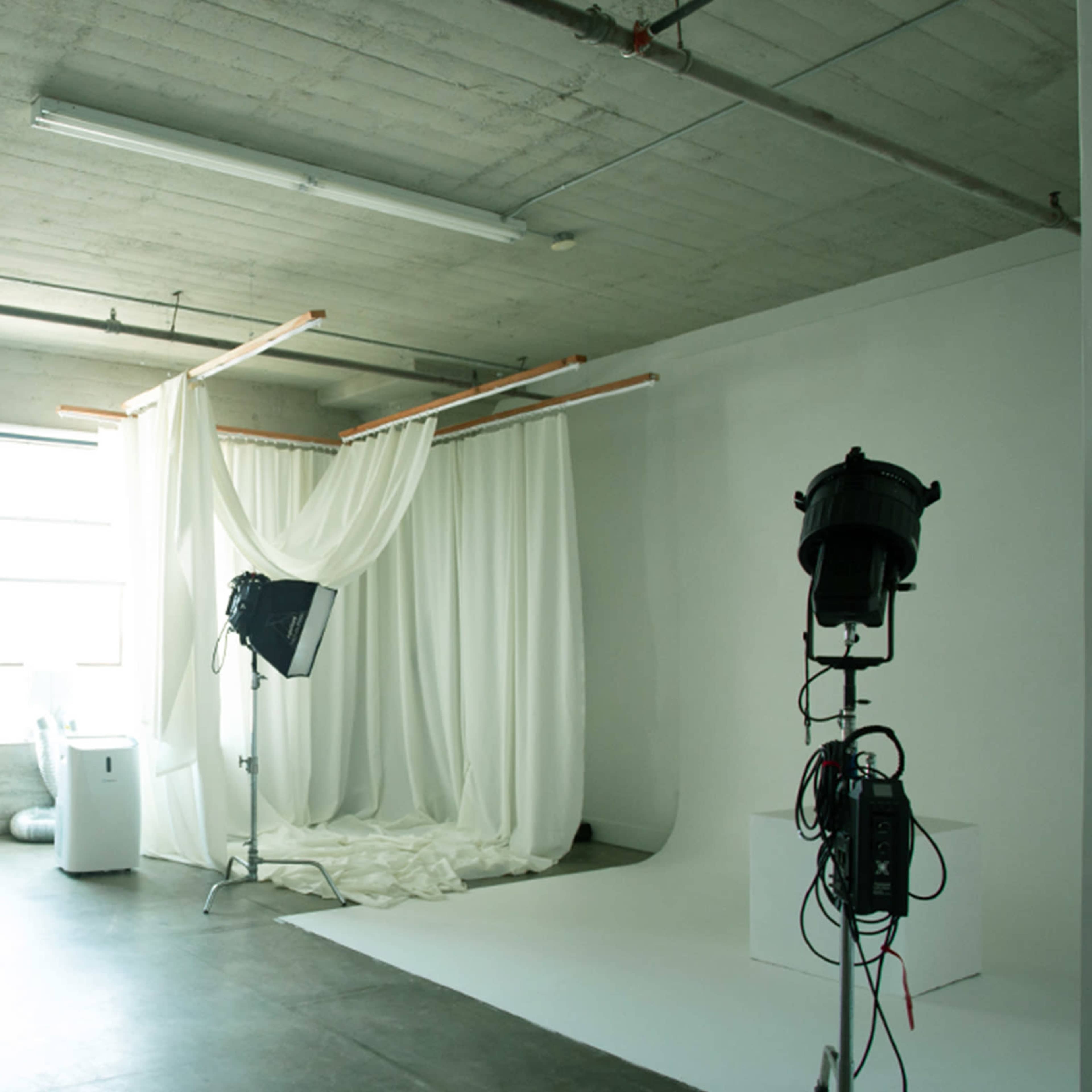A photography studio featuring soft white fabric draped in a corner, studio lighting equipment, and a plain white backdrop.
