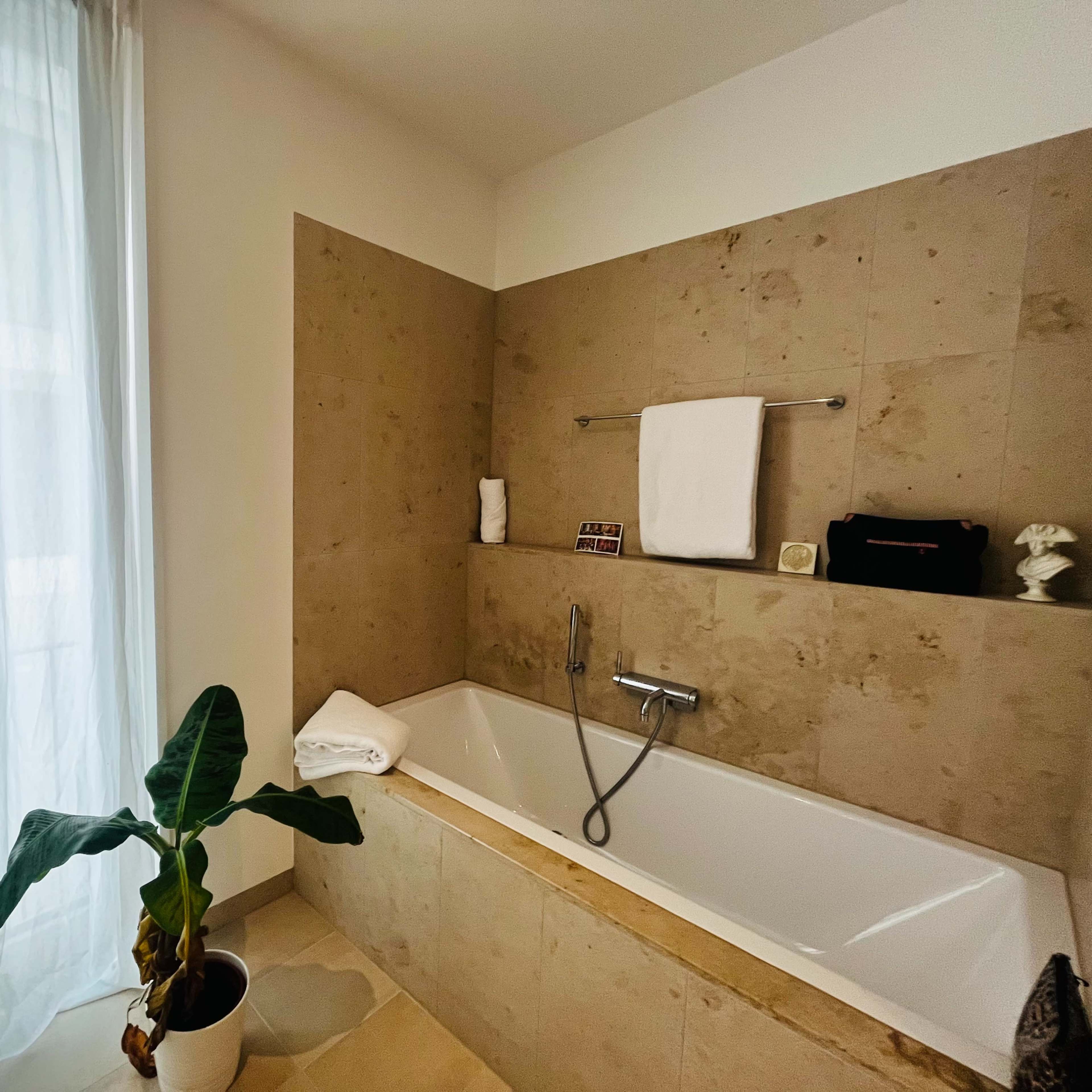 A bathroom features a white bathtub surrounded by beige stone walls, with a window covered by sheer curtains and a potted plant nearby.